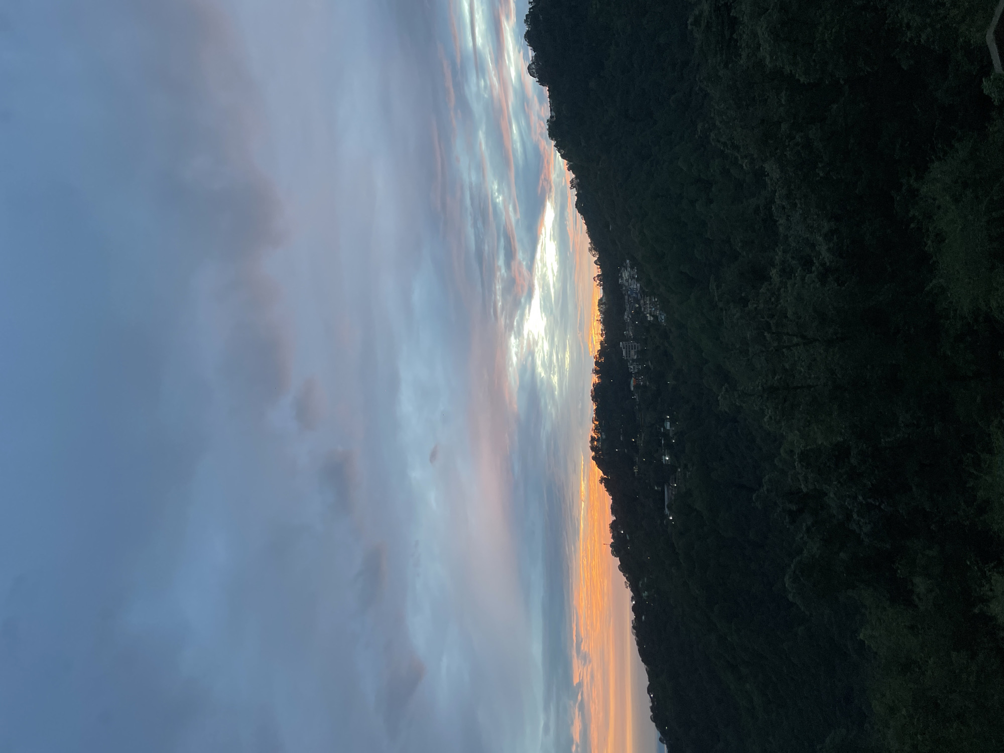 The image captures a serene sunset over a hillside landscape in Dharamkot, India. The sky is a canvas of muted blues and grays, streaked with soft hues of orange and pink from the setting sun. Clouds are scattered across the sky, with a bright band of light peeking through the horizon.
The hillside is densely covered with dark green trees, creating a rich, textured foreground. Scattered amongst the trees, particularly on the upper slopes, are clusters of buildings illuminated by small lights, indicating a settlement nestled within the natural environment. The architecture of these buildings is not clearly discernible due to the distance and lighting conditions.
The overall atmosphere of the image is tranquil and picturesque, emphasizing the beauty of nature and the quietude of the location as daylight fades.
