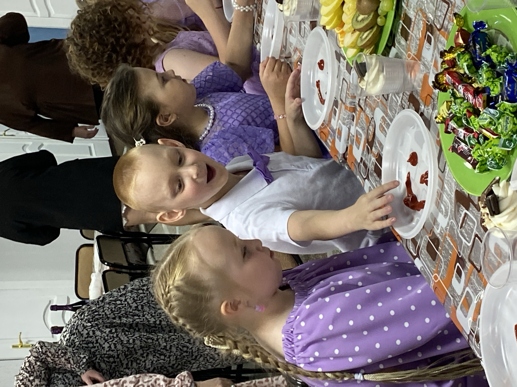 A horizontally-oriented photograph depicts a group of children at a table, presumably in Noviy, Russia. The children, ranging in age, are dressed in semi-formal attire, with one girl wearing a lilac dress with white polka dots. A boy is in a white shirt with a lilac bowtie. Another girl is in a lilac dress with a pearl necklace. A table is laid out with plates, food items like fruit slices, a container with a white creamy substance, and a dish full of wrapped candies. Some plates have small dollops of a red sauce in a smiley face pattern. The table has a patterned tablecloth, primarily in shades of brown, white and orange. In the background, part of an adult figure in brown and another in black are visible. Several chairs with dark frames and light-colored seats are situated behind the people.