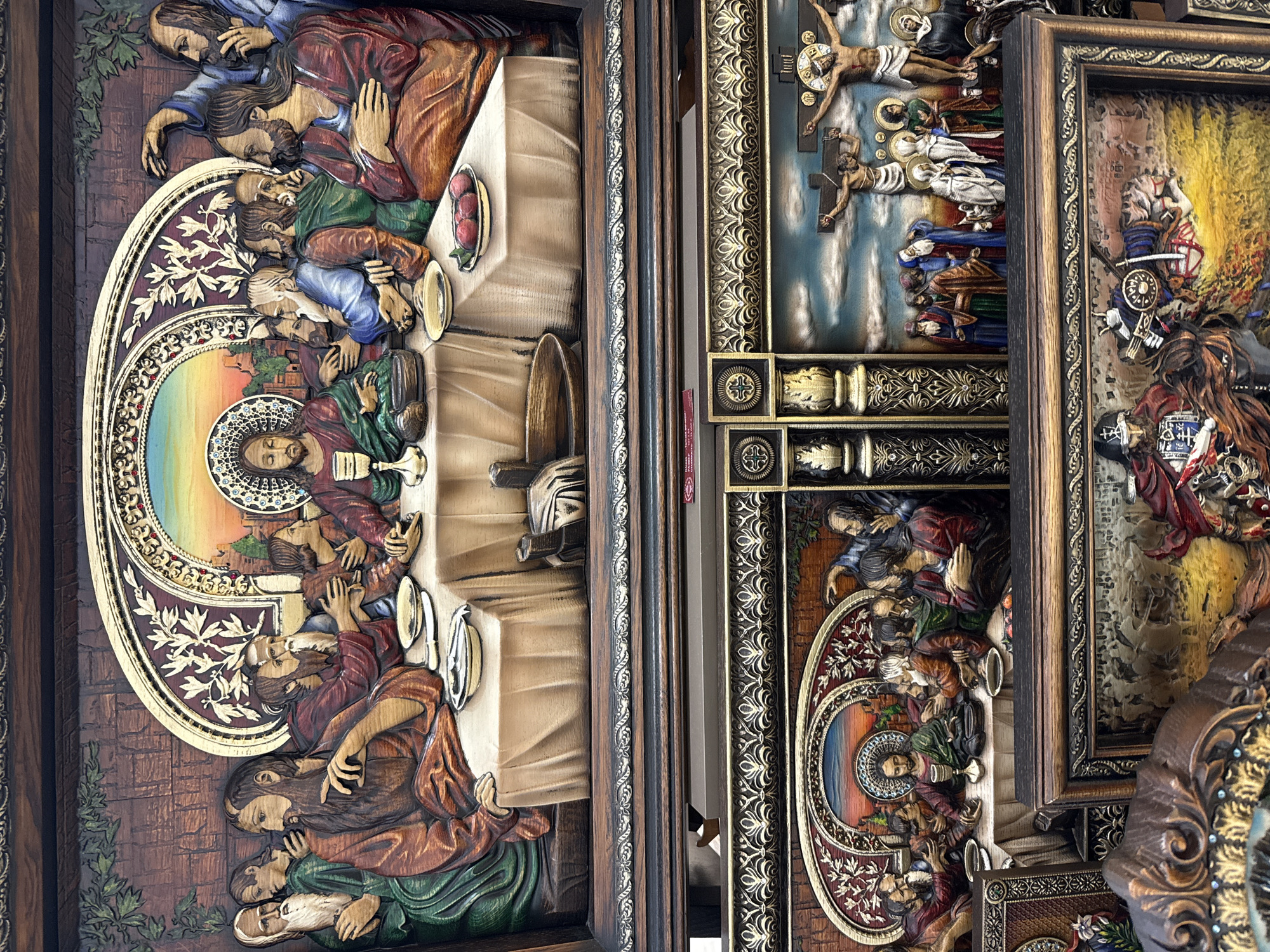 The image displays a diptych of intricately carved wooden religious scenes. The top panel depicts "The Last Supper," with Jesus at the center, surrounded by his twelve apostles seated at a long table covered with a tablecloth. The figures are rendered in relief with warm tones of wood and subtle painted details. Behind the table, a colorful, arched backdrop features a sunset sky over a city. The bottom panel presents two scenes. The left side shows another depiction of "The Last Supper," with Jesus and his disciples. The right side depicts the Crucifixion of Jesus Christ, with two other figures on crosses beside him and a crowd of onlookers below. The carvings are detailed, with elements like the texture of clothing, facial features, and the sky rendered with care. Both panels are framed by ornate, dark wood borders with decorative carvings. The overall presentation suggests a religious artifact or display of artwork.