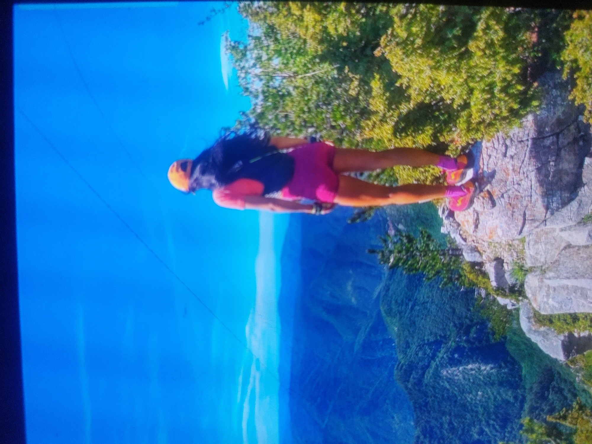 A person stands on the edge of a rocky cliff overlooking a deep valley. The person is wearing a helmet, a black and pink shirt, pink shorts, and pink sneakers. The cliff is covered in green vegetation. The valley is filled with trees and the sky is a bright blue. The photo was taken in Tsarevo, Bulgaria.
