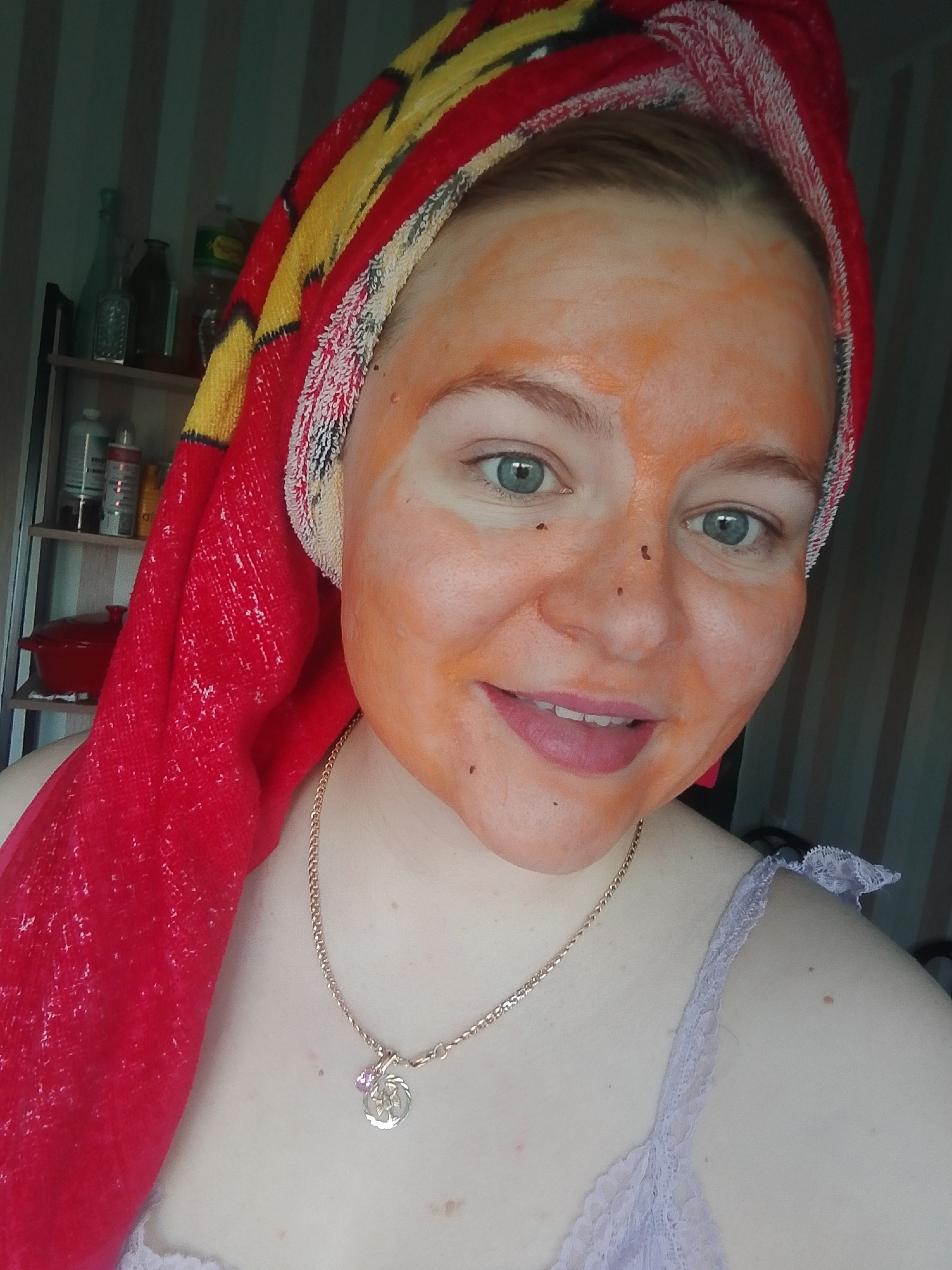 A woman with a face mask is smiling at the camera. She has light skin, blue eyes, and is wearing a red and yellow towel wrapped around her head. A gold chain necklace with a pendant is around her neck. She is wearing a light purple top. Behind her is a shelf with various bottles and jars. To the right, a striped window covering is visible. The location is not discernible as being in Izhevsk, Russia. There are no elements in the image which allow a location determination within the city of Izhevsk, Russia.