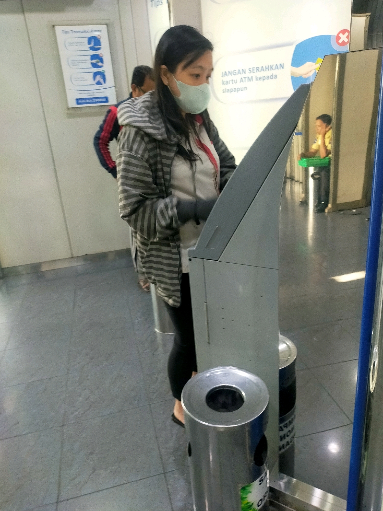 In a public indoor area, possibly a transit station or a shopping mall, a woman wearing a light blue face mask and a striped hooded jacket stands in front of a metallic kiosk or ticket machine. Her left hand is visible, wearing a dark glove. Behind her, a portion of another person in a dark jacket with red and blue accents is visible, as is a poster with text in Indonesian and graphic icons. To the right of the kiosk, a red "X" symbol is visible within a blue circle on a poster. Reflections in the metallic surface of the kiosk reveal a person in a yellow shirt sitting down, and cylindrical trash receptacles. The floor is tiled with large, grey square tiles. The overall impression is a functional, modern, indoor public space.