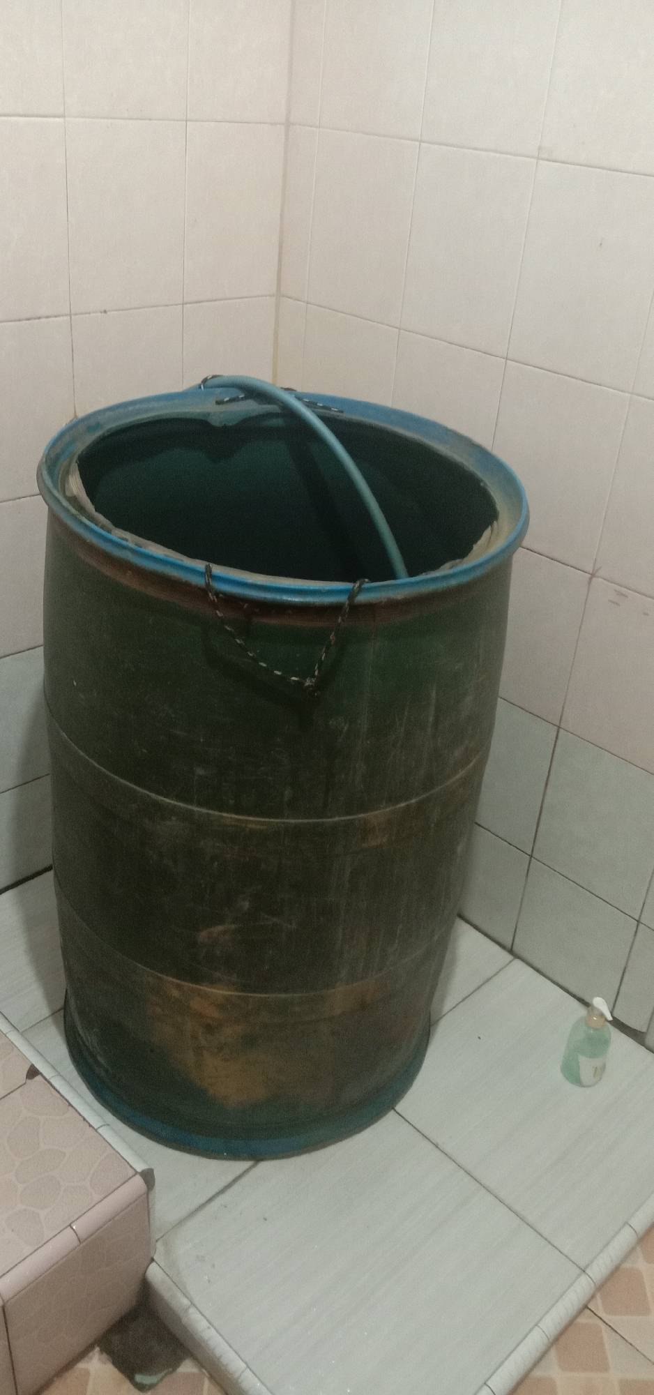 In a tiled corner of what appears to be a bathroom or utility area in Kendari, Indonesia, a large, dark green plastic barrel with a blue rim stands prominently. The barrel is cylindrical and shows signs of wear, with horizontal ridges circling its body. A light blue hose is draped over the top edge of the barrel, suggesting it might be used for filling or draining. Two small, dark metal chains are attached to the sides of the barrel's rim, possibly for handling or securing a lid. The floor is tiled with a light, neutral pattern, and a small, clear plastic bottle with a white pump dispenser, likely hand soap or sanitizer, is visible to the right of the barrel. The walls are also tiled with a similar light-colored pattern. The lighting suggests an indoor environment, possibly with overhead illumination. There are no people or animals present in the image.