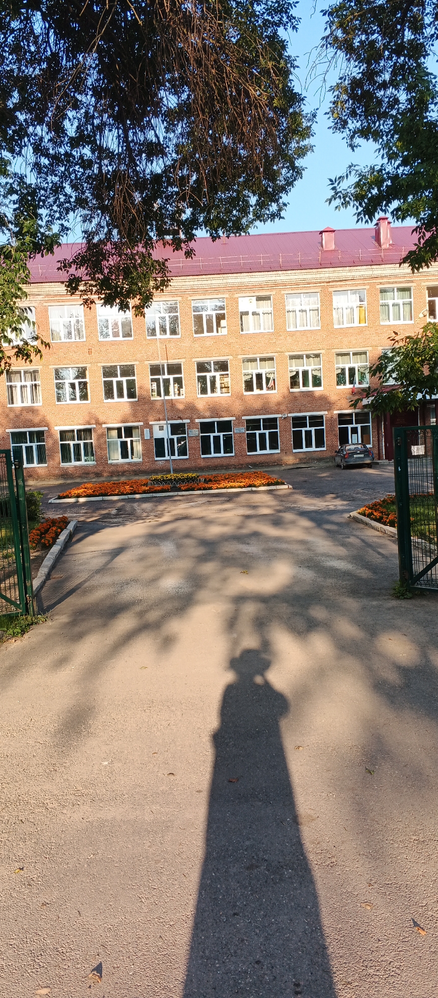 A wide shot shows a brick school building with a red roof, under a clear blue sky, partially obscured by tree branches. The building has multiple rows of windows, many of which are white-framed and reflect sunlight. In front of the building is a paved area with flower beds containing bright orange and yellow flowers. A fence made of green metal bars encloses part of the scene on the left, and a similar fence is visible on the right. A dark-colored car is parked on the right side of the paved area. The shadow of a person holding a camera, presumably the photographer, is cast prominently on the pavement in the foreground. The scene appears to be an entrance or courtyard area of the school. The context of Izhevsk, Russia, suggests this is likely a typical educational institution in the region.