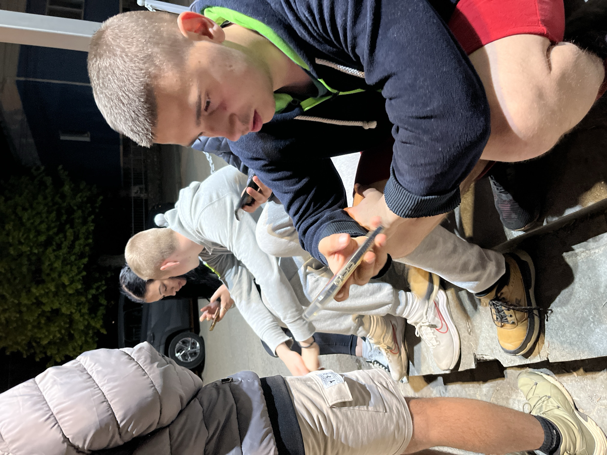 A group of young people are gathered on a concrete outdoor stairwell in what appears to be Karavelovo, Bulgaria. One young man wearing a dark hoodie with a bright green interior, red shorts, and dark sneakers is crouched and holds a smartphone in his right hand. He is looking down and appears to be focusing on something in front of him. Around him are three other people, two are looking at what the first one is holding. Another person on the left is looking at a smartphone. Each person is wearing different clothes and footwear. The surrounding environment is dark and lit by overhead lighting. In the background, there is a parked car and some trees.