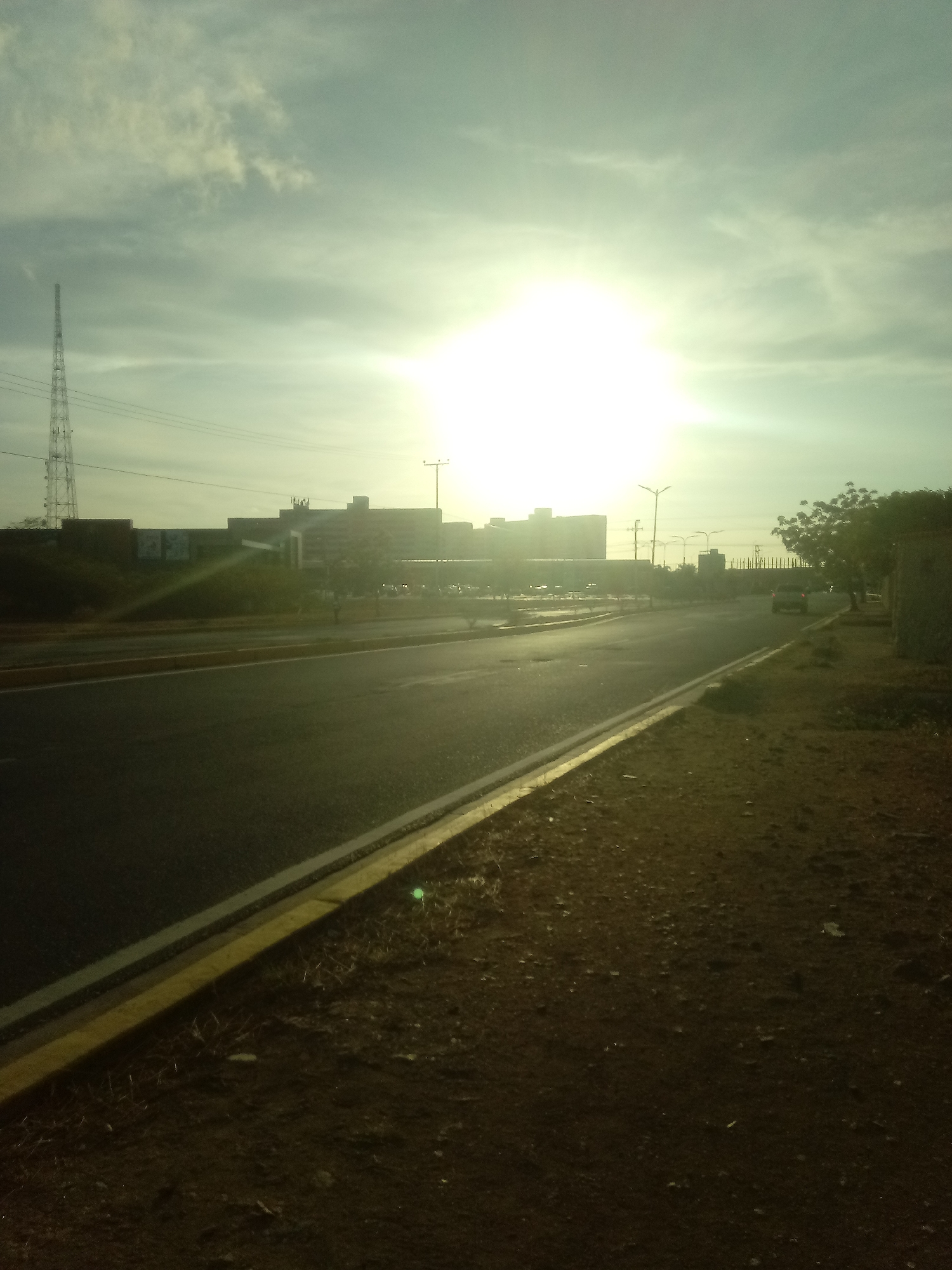 The image depicts an urban scene, possibly in Juan Griego, Venezuela, although without explicit verification, this is speculative. The perspective is from a low angle, likely ground level, alongside a road. The road is asphalt and features a yellow demarcation line. The area beside the road appears to be unpaved and dusty.
In the background, a large building or complex is visible, illuminated by the bright sun, making it difficult to discern precise details. A communication tower stands to the left of the building. Power lines stretch across the upper portion of the frame. A vehicle is visible on the road in the distance. Trees are present on the right side of the frame. The sky is overcast with a bright sun.