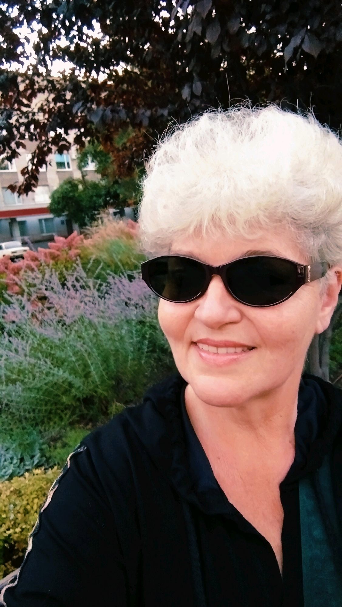 A medium shot photograph shows a woman with short, white, curly hair and dark sunglasses posing outdoors. She is wearing a black hooded jacket. The background consists of a blurred residential building, trees with dark leaves, and an area of ornamental grasses with pink and green hues. The setting appears to be a park or garden area in Horishni Plavni, Ukraine, as indicated by the residential architecture and landscaping typical of the region. The lighting suggests it is daytime, possibly late afternoon or early evening, with soft light filtering through the trees.