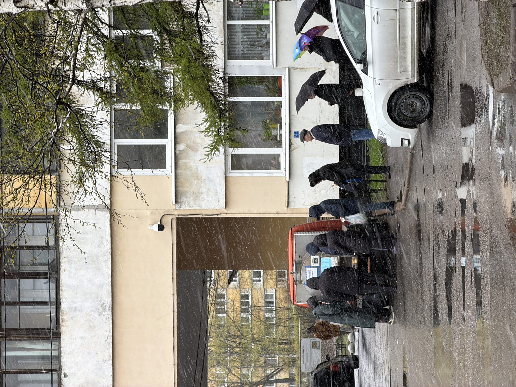 The image depicts a group of people lined up at an ATM in Saint Petersburg, Russia, during a period of snowfall. The ATM is red and silver, and the people are dressed in winter clothing and holding umbrellas to shield themselves from the snow. A white car is parked next to the ATM. The street is wet, reflecting the buildings and figures. The buildings in the background are light-colored and have multiple windows. A birch tree with light green leaves is visible in the upper left corner of the frame. A security camera is mounted on the building.