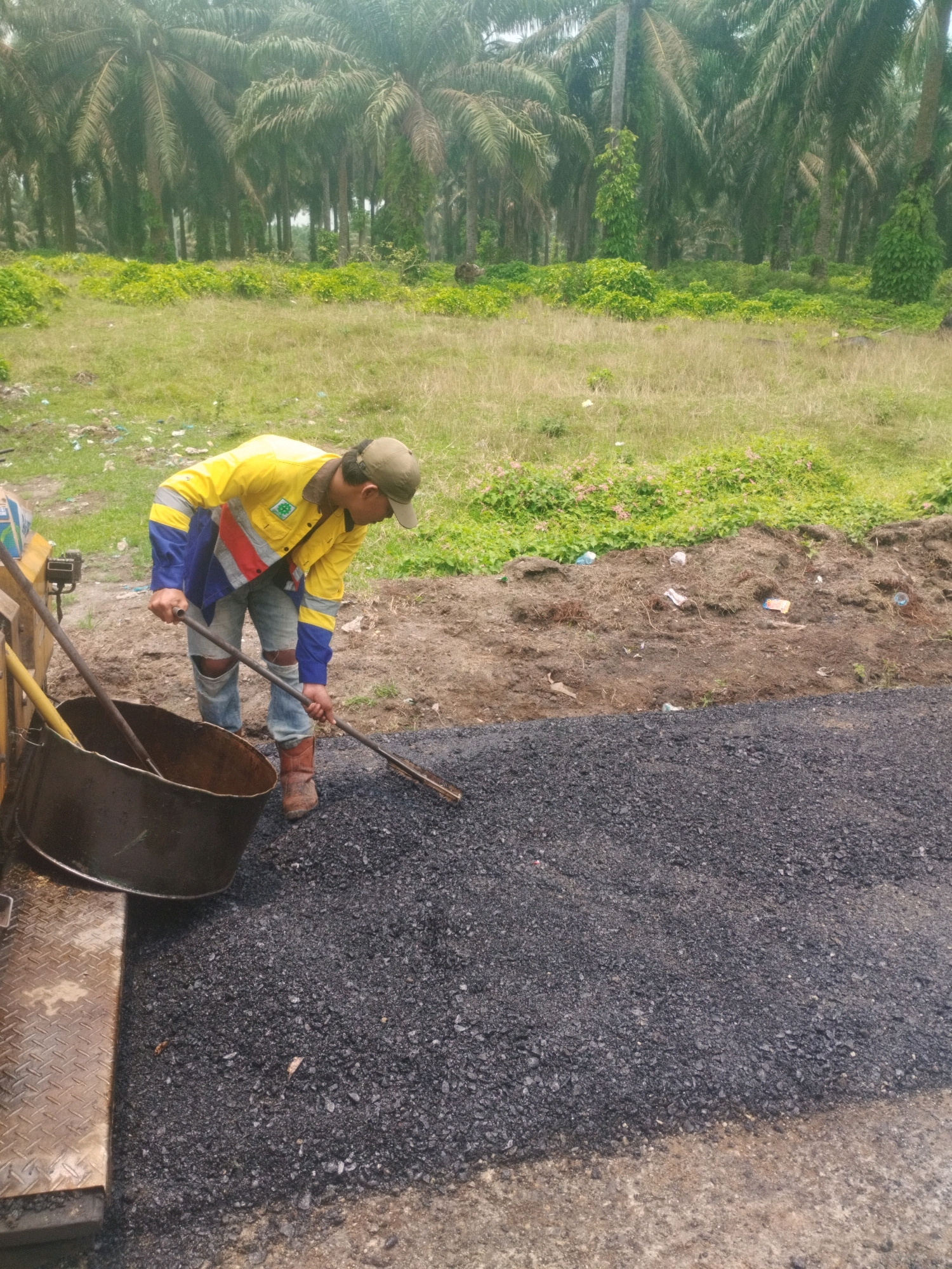 A person is spreading asphalt on a road. The person is wearing a yellow and blue long-sleeved shirt, jeans, and brown boots. They are also wearing a cap. The person is using a long-handled tool to spread the asphalt. There is a large black pot next to the person. In the background, there are palm trees and green vegetation. The road is made of black asphalt. The image appears to have been taken in the city of Pagarbosi, Indonesia.