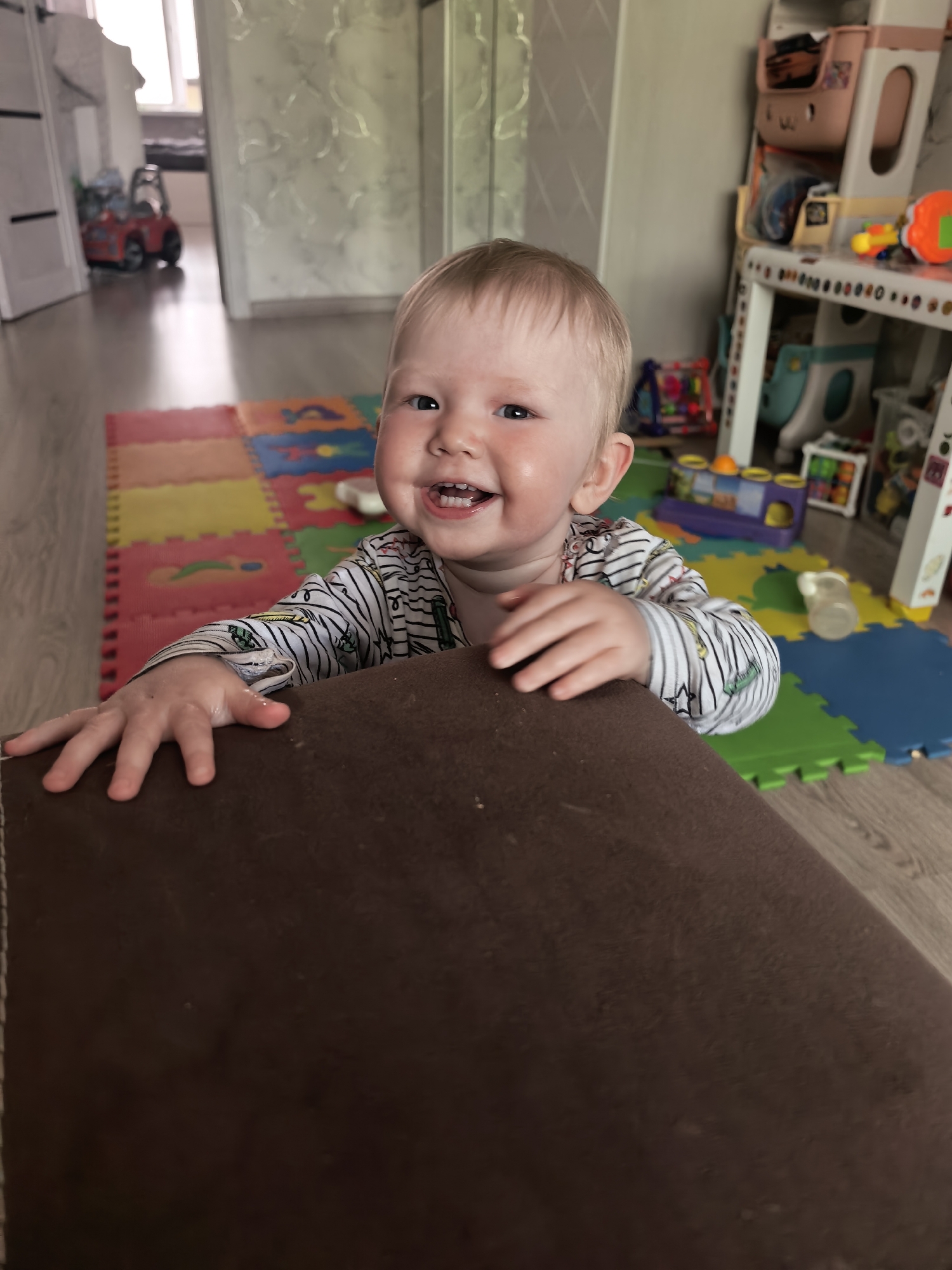 A fair-skinned infant with blond hair is standing behind a dark brown upholstered object. The child is smiling and has their hands on the object. The background includes a colorful play mat, toys, and furniture. The location is likely a domestic interior. It's not possible to confirm the location to be Sterlitamak, Russia based on the provided image.