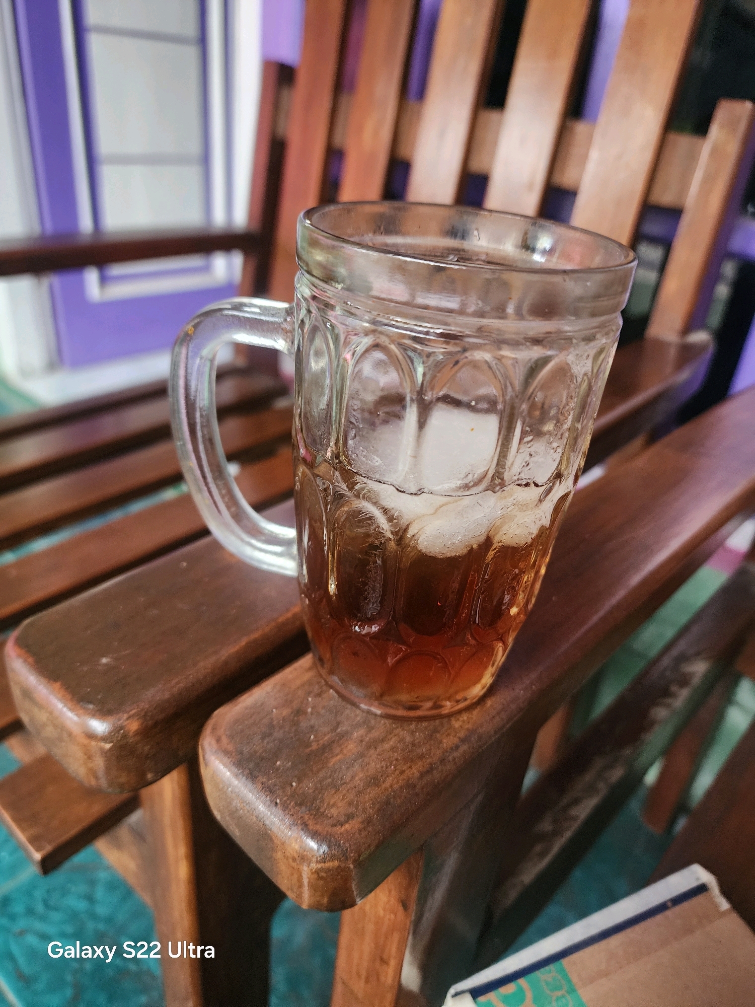 A close-up, eye-level shot features a clear glass mug filled with iced tea, positioned on the wooden slats of a chair. The mug is roughly half-full with a dark amber liquid and several ice cubes. The mug itself has a textured, ribbed design and a C-shaped handle. The wooden chair is made of dark brown wood with a slatted backrest visible in the background. The background also includes hints of purple and white, likely from a building or wall. The bottom left corner of the image displays the text "Galaxy S22 Ultra". The scene appears to be an outdoor setting, possibly a porch or patio.