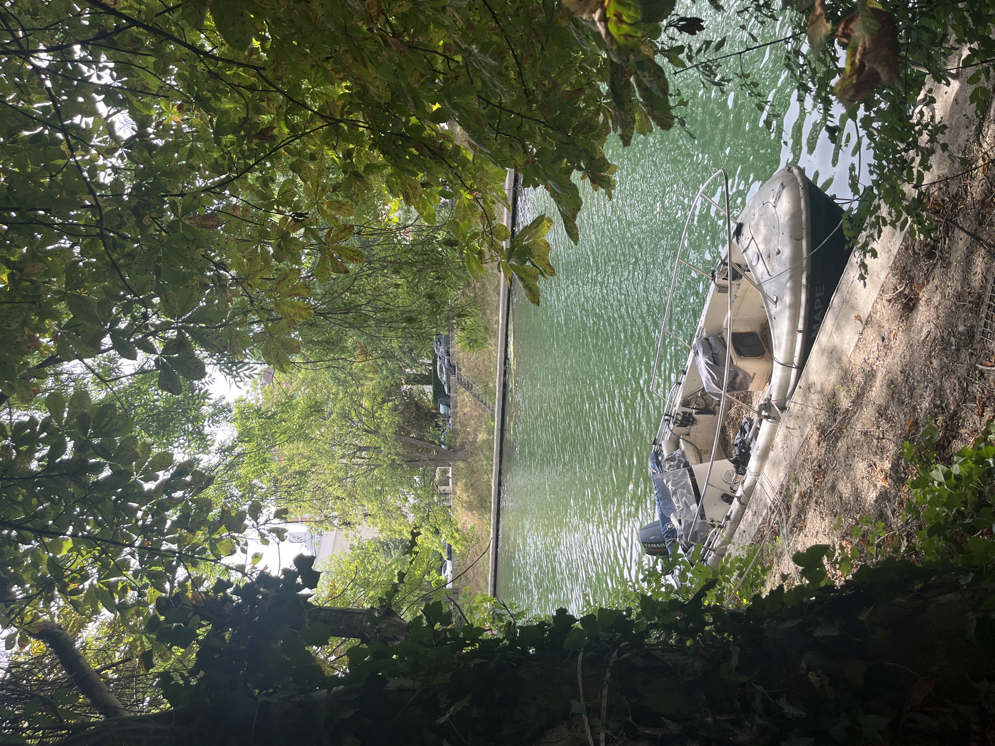 An inflatable boat with a Yamaha outboard motor is moored along the bank of a green canal in Joinville-le-Pont, France. The boat is situated on a concrete embankment, partially shaded by overhanging trees and foliage. The water reflects the sunlight, creating a dappled effect. In the background, across the canal, is a tree-lined street with parked cars and buildings. The scene is peaceful and appears to be a typical summer day.