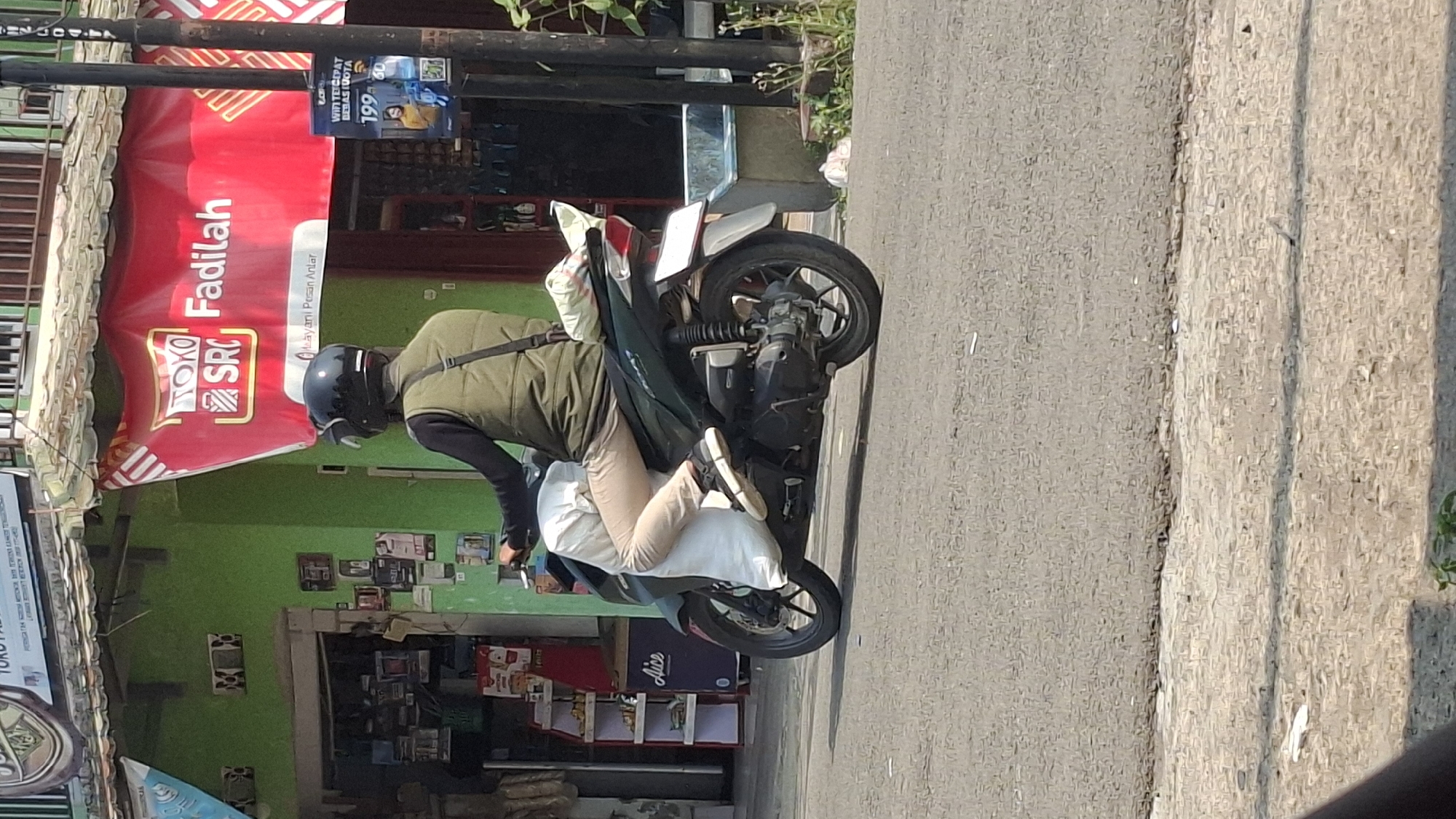 The image depicts a street scene in Cilaku, Indonesia, with a person riding a motorcycle. The rider is wearing a dark helmet and a light olive-green vest over a dark long-sleeved shirt. They are also wearing light-colored pants and dark sneakers. The motorcycle is dark green and is carrying two large white bags, one on the front of the rider and one behind. The background shows a building with a bright green facade and a large red banner advertising "TOKO SRC Fadilah". A street sign with "Layanan Pesan Antar" (Delivery Service) is also visible on the banner. To the right of the building, a utility pole with some advertisements is present, including one for "WIFI TERCEPAT" (Fastest WiFi) with a price of "199". The street itself is paved, and there is a sidewalk area with some vegetation and debris. The lighting suggests it is daytime, likely sunny.