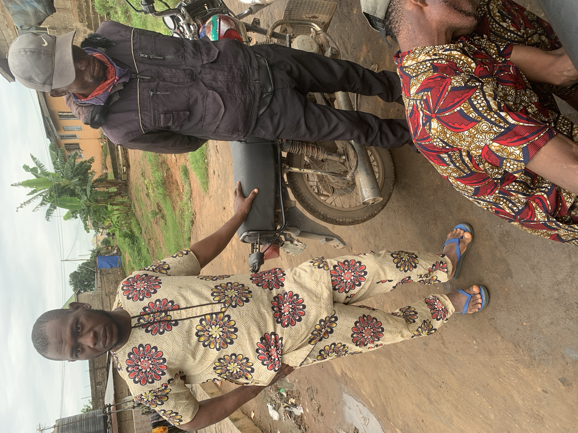 In Shagamu, Nigeria, two men and a motorcycle are visible. One man, wearing a patterned beige and red outfit with sandals, stands with his hands on his hips. Another man, dressed in a dark jacket, black pants, and a baseball cap, stands with his head bowed. A third individual, wearing a patterned red and blue shirt, is bent over on the ground. A motorcycle is parked nearby. The ground appears to be dirt. Buildings and vegetation are in the background.