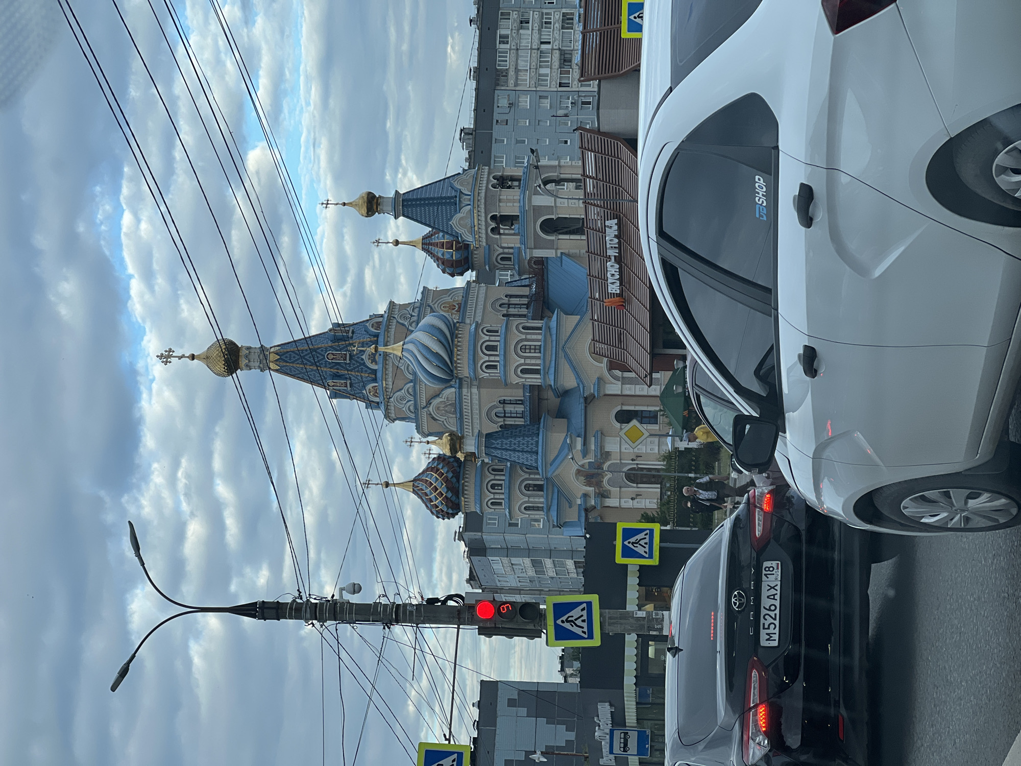 This image captures a street view in Izhevsk, Russia, likely during daylight given the sky and lighting. The scene is dominated by a prominent, ornately decorated Orthodox church with blue and gold domes. In the foreground, several cars are visible, stopped in traffic. A white car is closest to the camera, with a portion of its rear and side showing. Behind it, a black Toyota Camry is also stopped, its license plate clearly visible as "M526 AX 18".
Traffic lights are present, with a red light illuminated, indicating that the vehicles are currently stopped. Pedestrian crossing signs and other road signage are also visible. Overhead power lines crisscross the sky. In the background, beyond the church, modern apartment buildings can be seen.
A sign on the fence in front of the church reads "ВКУСНО – И ТОЧКА", which translates to "Tasty – and that's it" or "Tasty – That's the spot," suggesting a nearby food establishment. A sticker on the white car's window displays "DB SHOP". The weather appears to be overcast, with clouds filling the sky. The overall impression is a typical urban street scene in Russia, with a mix of historical architecture, modern infrastructure, and everyday traffic. No specific emotions are discernible, but the scene suggests a moment of pause in the flow of city life.