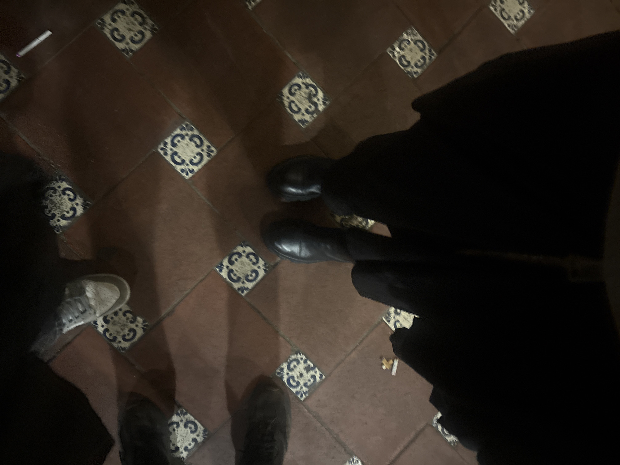 The image shows a tiled floor with four pairs of feet. Three pairs wear closed-toe shoes: one gray sneaker, one black boot, and one black shoe. A cigarette butt is visible on the floor. The location is likely Mexico City, Mexico, based on the style of the tiles.