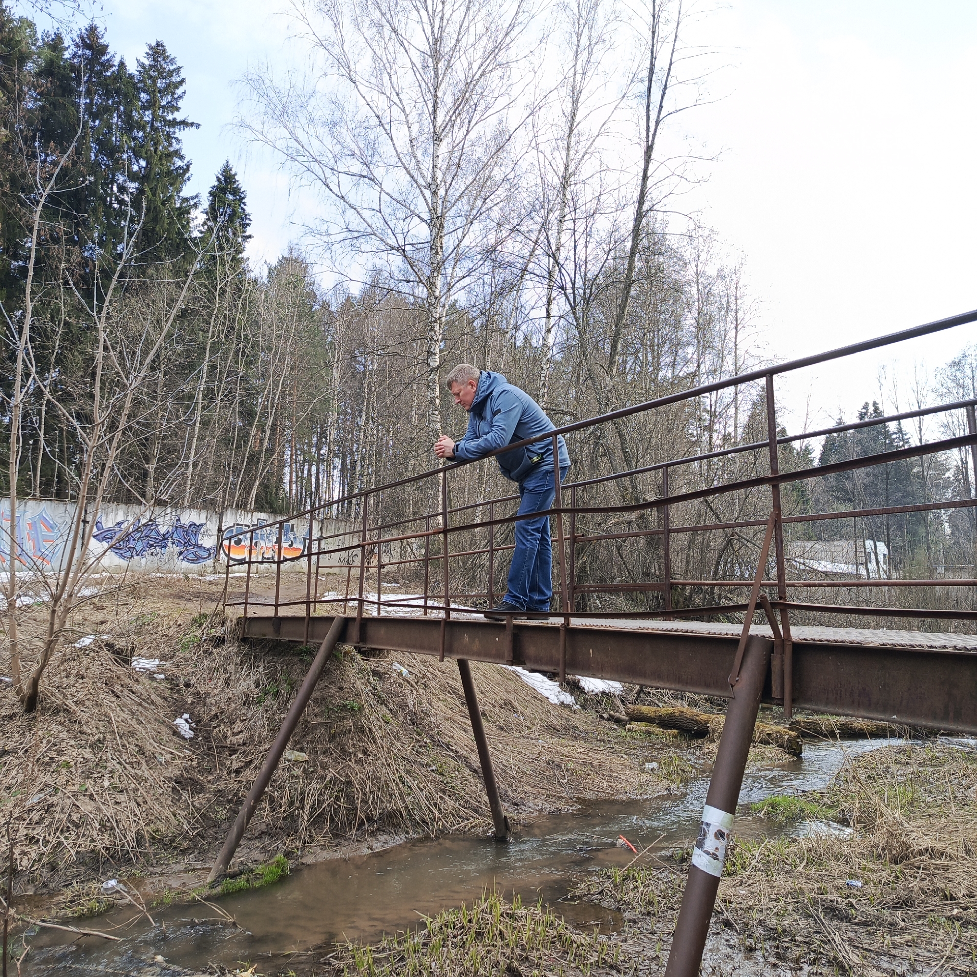 A man stands on a rusted metal bridge over a small stream in Chaikovsky, Russia. The bridge has a railing. The man, dressed in a blue jacket and jeans, leans on the railing. The environment includes early spring vegetation and a wall with graffiti. Trees are bare, and the ground is muddy.