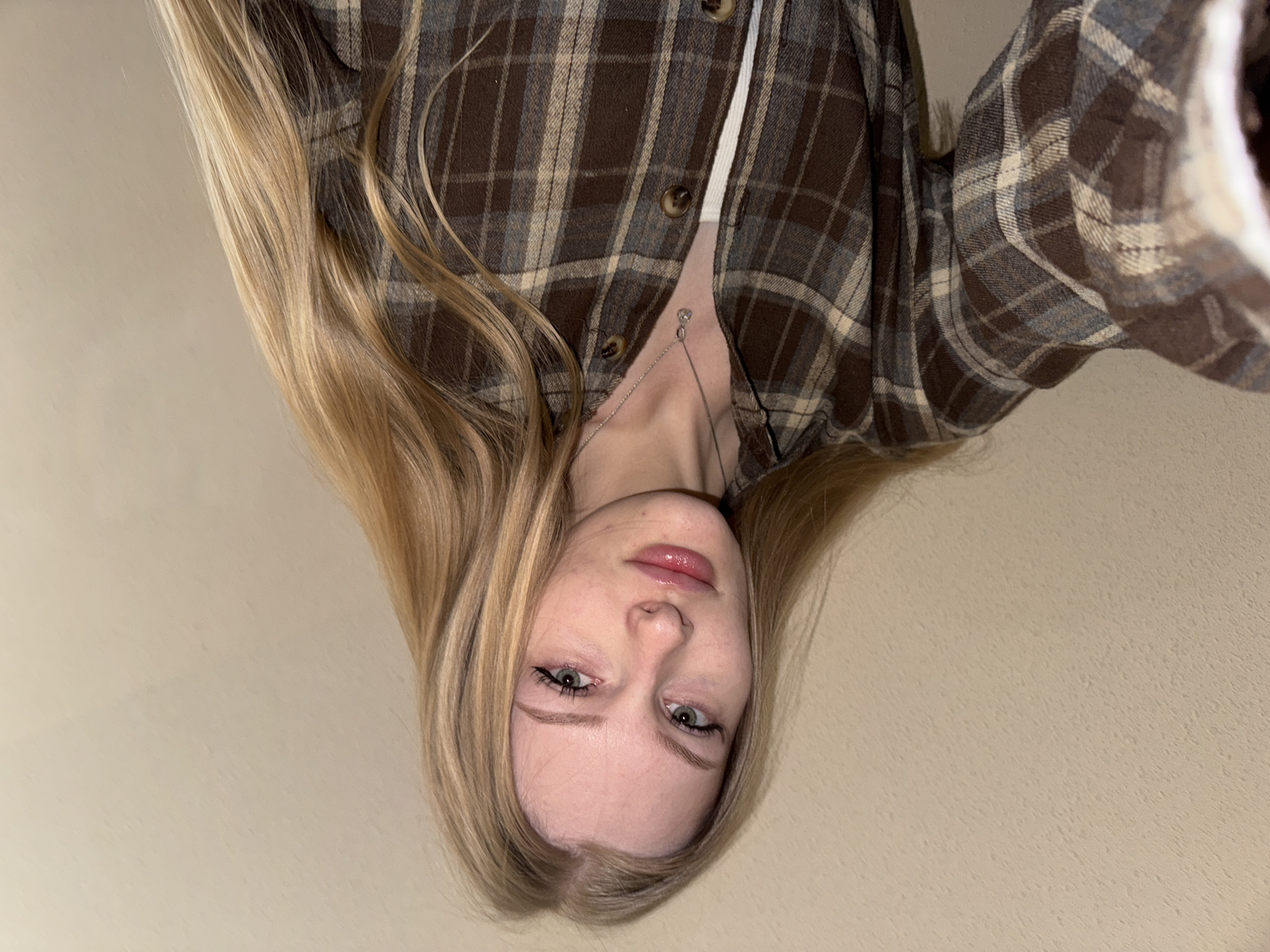 A young woman with long blonde hair and fair skin is the primary subject. She's wearing a brown and tan plaid shirt over a white top, with a silver necklace visible. The background appears to be a plain, light-colored wall. The image appears to have been taken from an overhead angle, resulting in a slightly distorted perspective. There is no clear indication of location. The city of Perm, Russia, is not discernible within the image.