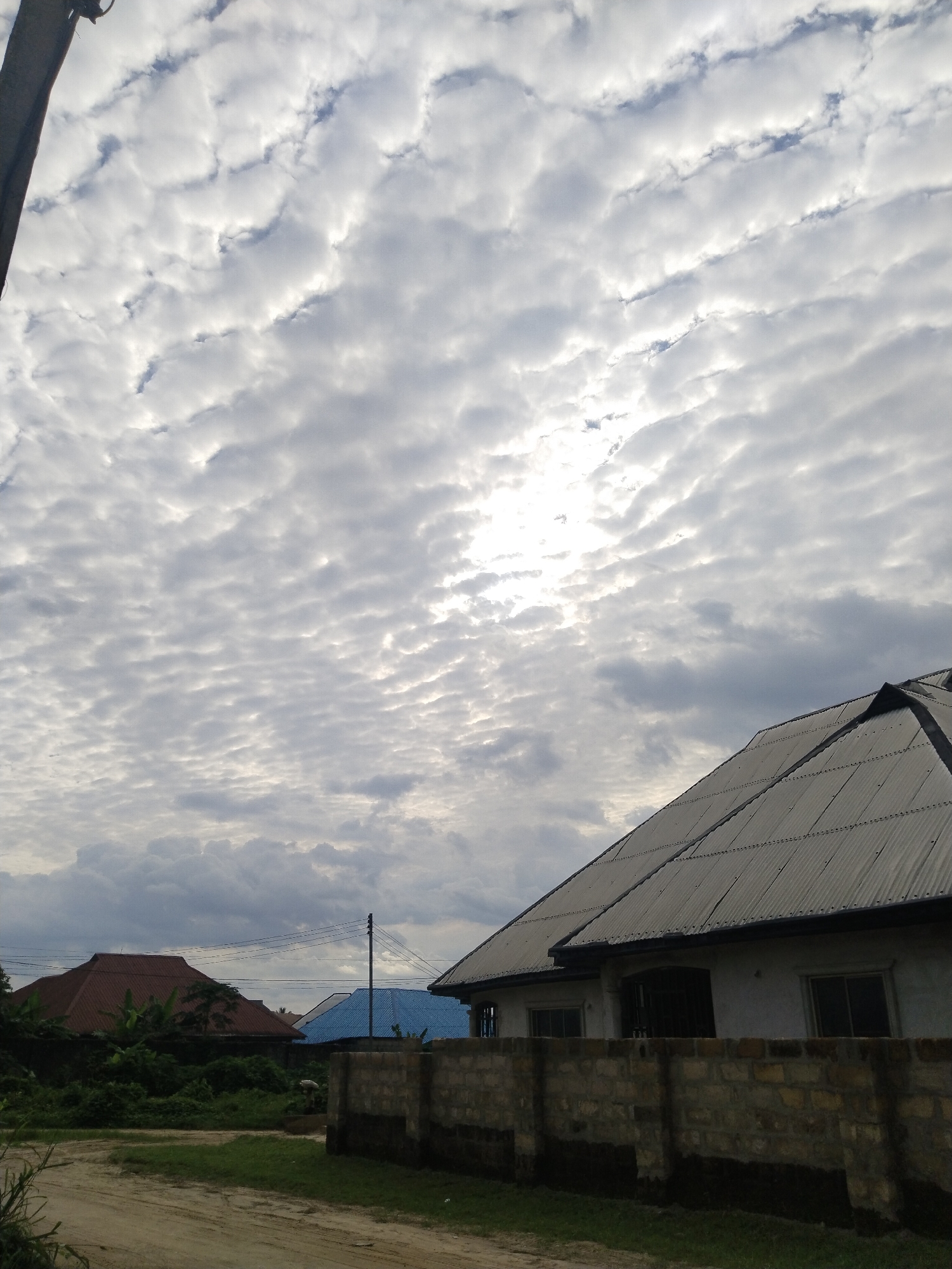 The image depicts a cloudy sky above a residential area in Uyo, Nigeria. The sky is filled with altocumulus clouds, creating a textured, slightly overcast appearance. Below the sky, the scene includes houses with corrugated metal roofs. One house is particularly prominent with a light-colored wall and a brick fence. In the background, there are glimpses of other houses with roofs painted in red and blue. Green vegetation is interspersed throughout the scene, suggesting a somewhat rural or suburban environment. The foreground shows a dirt road or path, indicating the location's relatively undeveloped nature. A utility pole is visible on the left. There is no visible human activity or interaction.