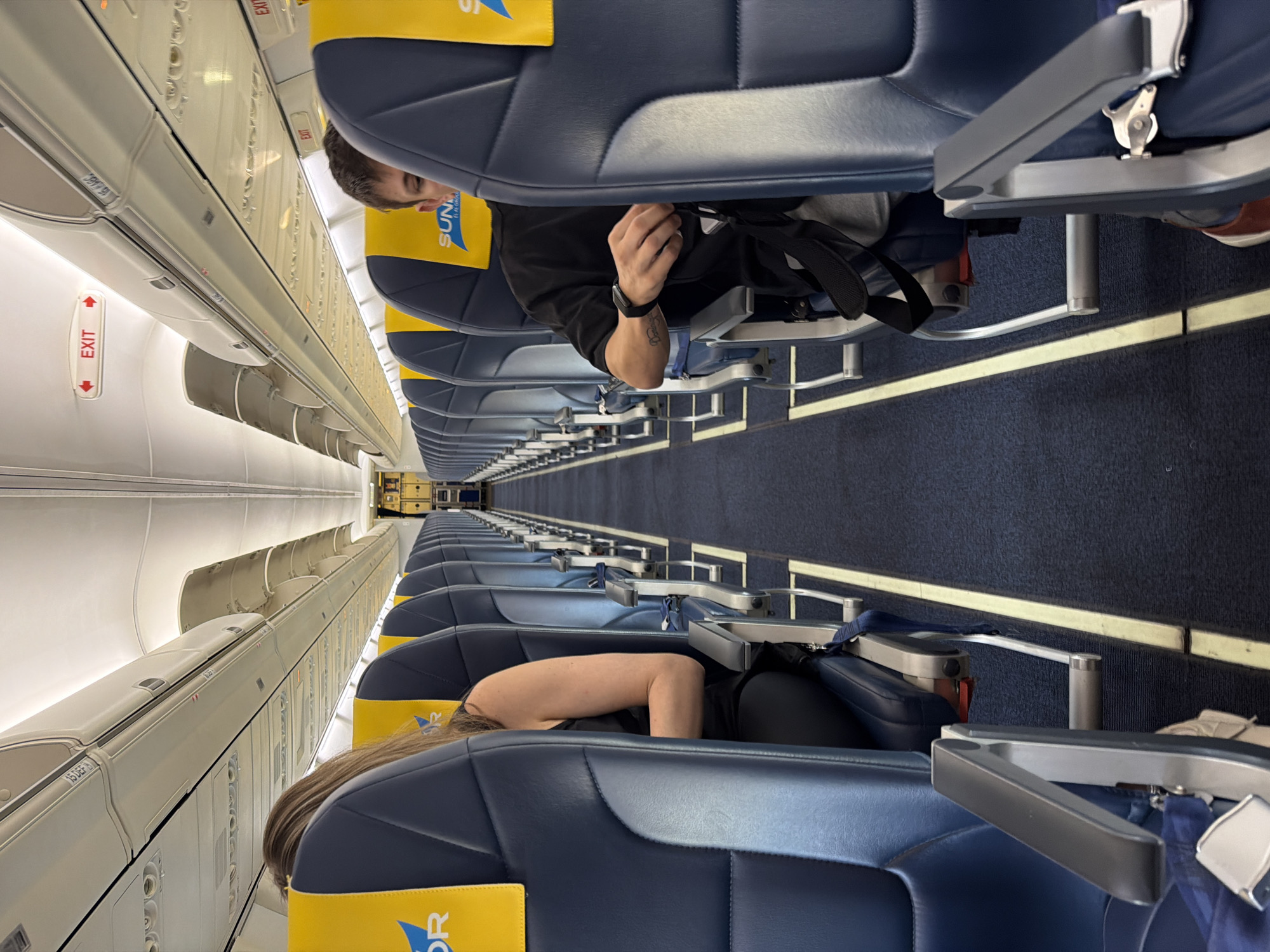 An interior shot shows the aisle of an airplane cabin, looking forward. The seats, upholstered in navy blue with yellow headrests, are arranged in rows on both sides. A woman with long brown hair is seated on the left, and a man with short brown hair is seated on the right. Both individuals appear to be passengers. The man is wearing a black t-shirt and a watch on his left wrist. The woman is wearing a black sleeveless top. Overhead compartments are visible along the ceiling. Red "EXIT" signs with arrows are mounted on the ceiling of the cabin. The floor is covered with a dark blue carpet with yellow markings. The image appears to have been taken from a standing position within the aisle. The location is Unknown, Israel.