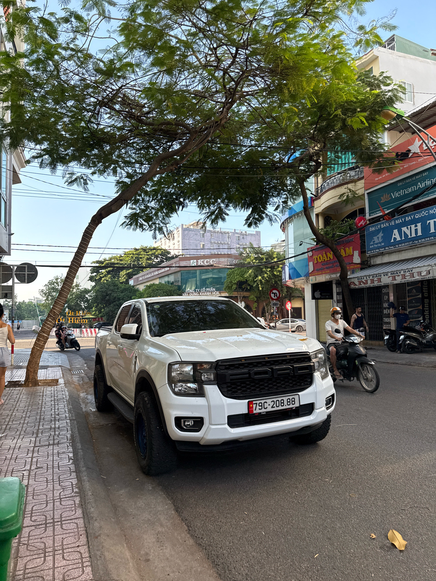 A white Ford pickup truck is parked on the right side of a paved city street in Nha Trang, Vietnam. The truck is angled towards the viewer, with its front bumper and grille clearly visible. The license plate reads "79C-208.88" with a Vietnamese flag emblem. A tree with lush green foliage overhangs the street and the truck, casting shadows. In the background, buildings with colorful facades line the street, some displaying signs with Vietnamese text. A man on a motorcycle is riding past the truck on the right side of the road. Further down the street, another motorcycle is parked. On the left side of the image, a sidewalk is visible, with a person walking away from the camera. Power lines crisscross above the scene.