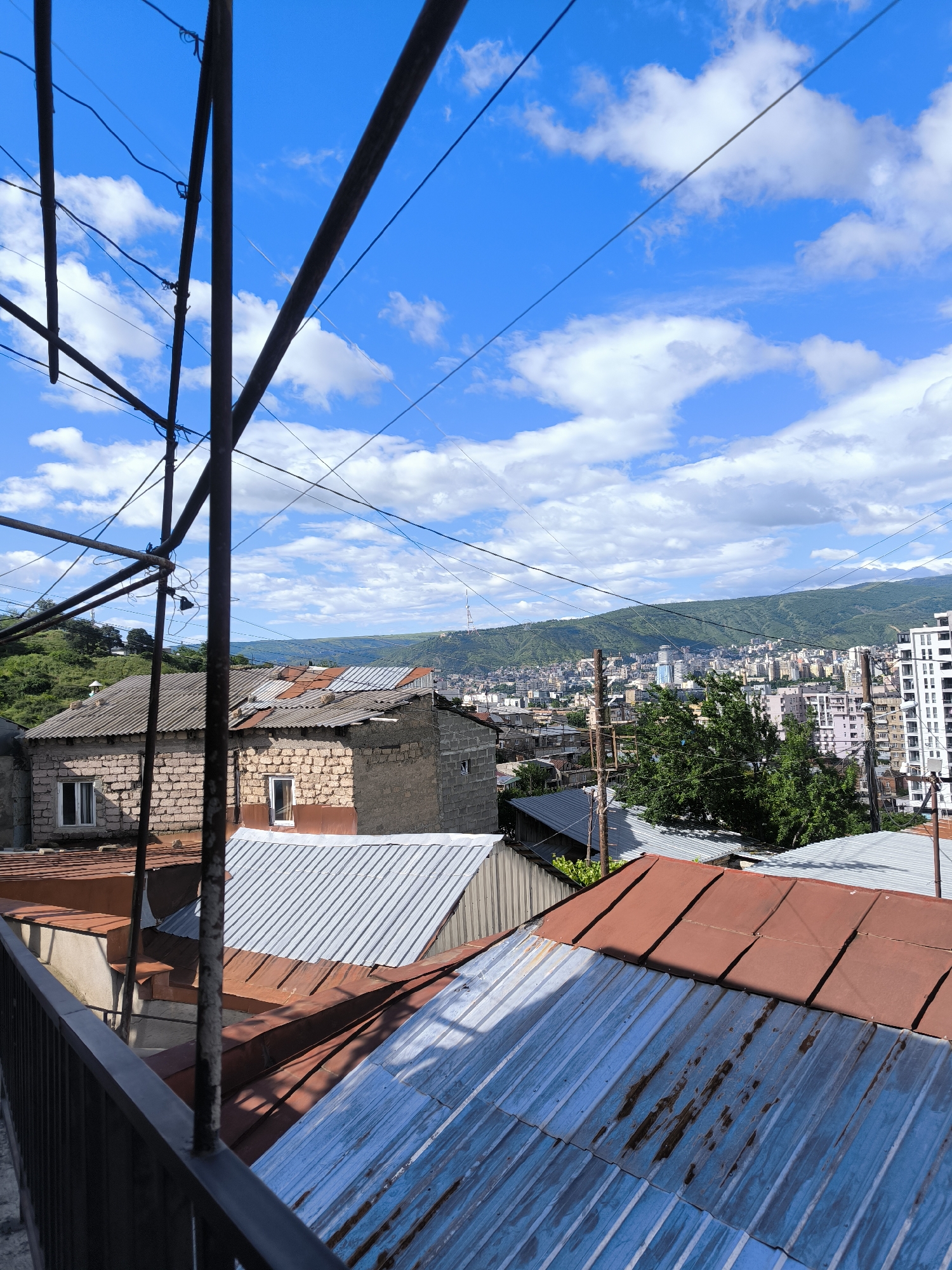 The image captures a view from an elevated position, likely a balcony or rooftop, overlooking a section of Tbilisi, Georgia. In the foreground, various rooftops made of corrugated metal sheets dominate, exhibiting a mix of rust-colored and silver/blue tones. A metal railing of a balcony is visible on the lower left. Electrical wires crisscross the sky, and metal support structures are present on the left side of the frame.
In the middle ground, there are older, brick or stone buildings with tiled roofs. Beyond these buildings, the cityscape stretches out, revealing an array of residential and commercial structures. A tree is visible amid the city skyline.
The background showcases a mountain range under a bright blue sky filled with scattered white clouds. The scene seems to be captured during the daytime. The architectural styles are typical of older residential areas in Tbilisi.