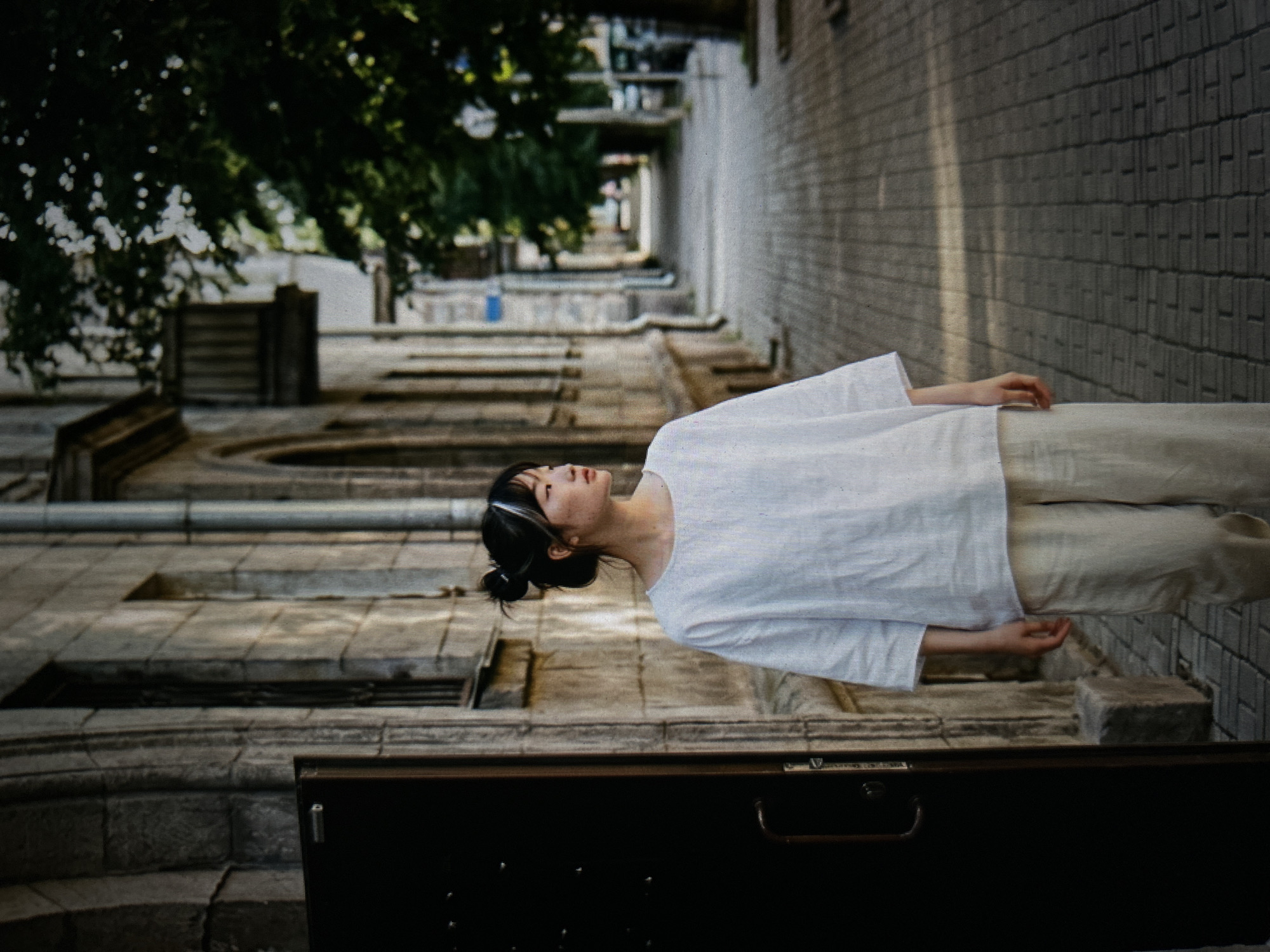 A young East Asian woman with her dark hair pulled up in a bun stands on a paved sidewalk in front of an old stone building. She is wearing a loose-fitting white linen shirt and light-colored linen pants. The woman is looking to her left, away from the camera. The building behind her has a dark-colored door on the left side of the frame, and a row of windows running along its facade. To the right of the woman, the sidewalk continues into the distance, lined with trees. The lighting suggests it is daytime, with some dappled sunlight on the pavement.