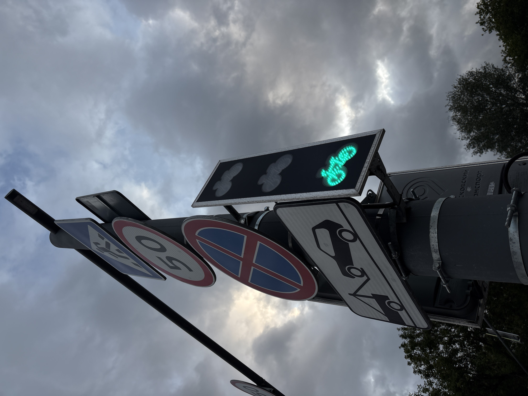 The image shows a traffic light signal pole with multiple signs and a traffic light. The traffic light displays a green signal in the shape of a pedestrian, indicating that pedestrians can cross. The pole is located in Moscow, Russia.
The signs visible on the pole include:
* A blue square sign with a white pedestrian symbol, indicating a pedestrian crossing.
* A red and white circular sign with "50" in black, indicating a speed limit of 50 km/h.
* A blue and red circular sign with a red cross, indicating no stopping or parking.
* A white rectangular sign with a black silhouette of a tow truck towing a car, indicating that vehicles will be towed.
The traffic light itself is a vertical panel with three illuminated symbols. The bottom symbol, which is illuminated green, depicts a stylized figure of a person walking, signaling for pedestrians to cross. The two upper symbols, which are unlit, appear to be cloud-like shapes.
The background consists of a cloudy sky, with some patches of brighter sky visible, suggesting it might be daytime with overcast conditions. Trees are visible in the lower left and right portions of the image. The pole itself is dark gray and attached to a larger cylindrical structure by metal bands. There is also some Cyrillic text visible on the larger structure below the signs.