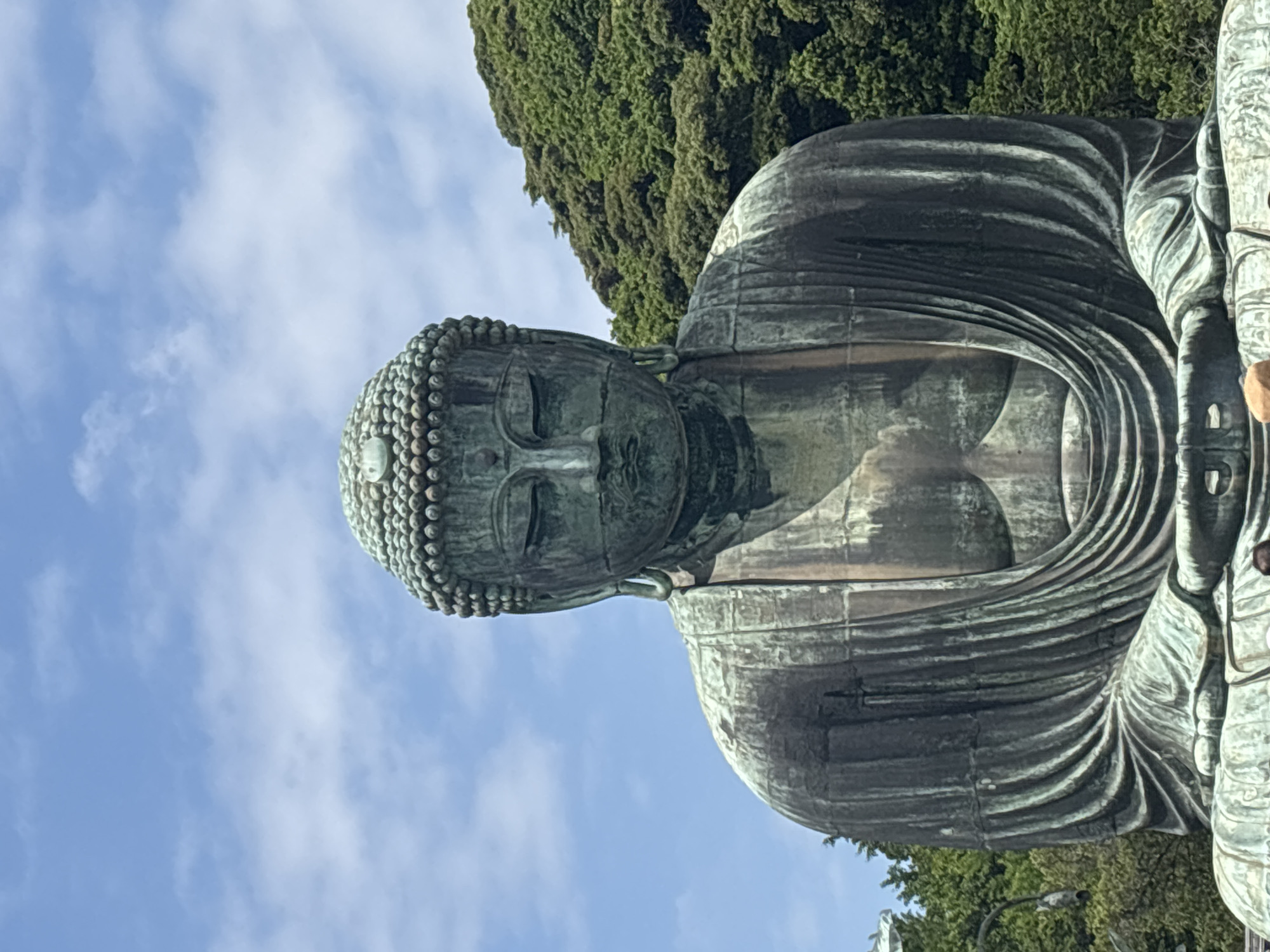 The image captures a frontal, medium shot of the Great Buddha of Kamakura, a colossal bronze statue situated in Kotoku-in Temple, Kamakura, Japan. The statue is depicted seated in a meditative pose, with its eyes closed and a serene expression. The intricate details of its robes and the curls of its hair are visible. Behind the Buddha, a lush green hillside covered in trees forms the backdrop, with a clear blue sky dotted with white clouds above. The statue is weathered, showing a greenish patina typical of aged bronze. The lower portion of the image reveals the base of the statue and hints at the presence of other visitors, though they are out of focus and partially obscured. The lighting suggests it is daytime, with sunlight illuminating the scene.