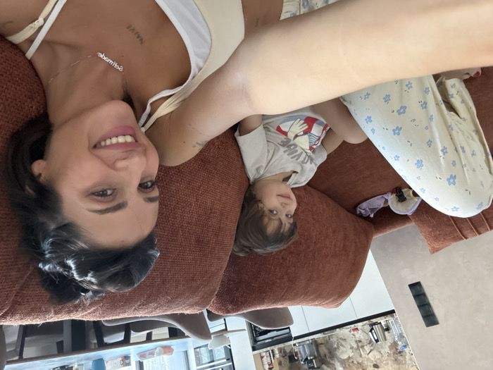 An indoor selfie captures an adult female and a young child seated on a terracotta-colored sofa within a residence in Khimki, Russia. The woman, positioned to the right and in the foreground, extends her right arm to hold the camera. She has dark hair styled in an updo and smiles towards the camera. She wears a light-colored tank top, a silver necklace with a word (possibly "Naomi" reversed), and a tattoo displaying "NAIVE" on her upper chest. Her left leg is partially visible, clad in light-colored pajama pants with a blue floral pattern. To her left, a young child with dark, curly hair sits on the sofa, also smiling towards the camera. The child wears a light-colored t-shirt featuring a "GOOD VIBE" graphic. Behind the child, on the sofa, are several folded fabric items. The background reveals an open-plan kitchen and dining area. The kitchen features light-colored countertops, dark stone-patterned upper cabinets, a silver electric kettle, and a water bottle. A textured, light-colored wall with a black multi-segment control panel is visible. Beyond the kitchen counter, a dark-colored dining table with matching chairs is present.