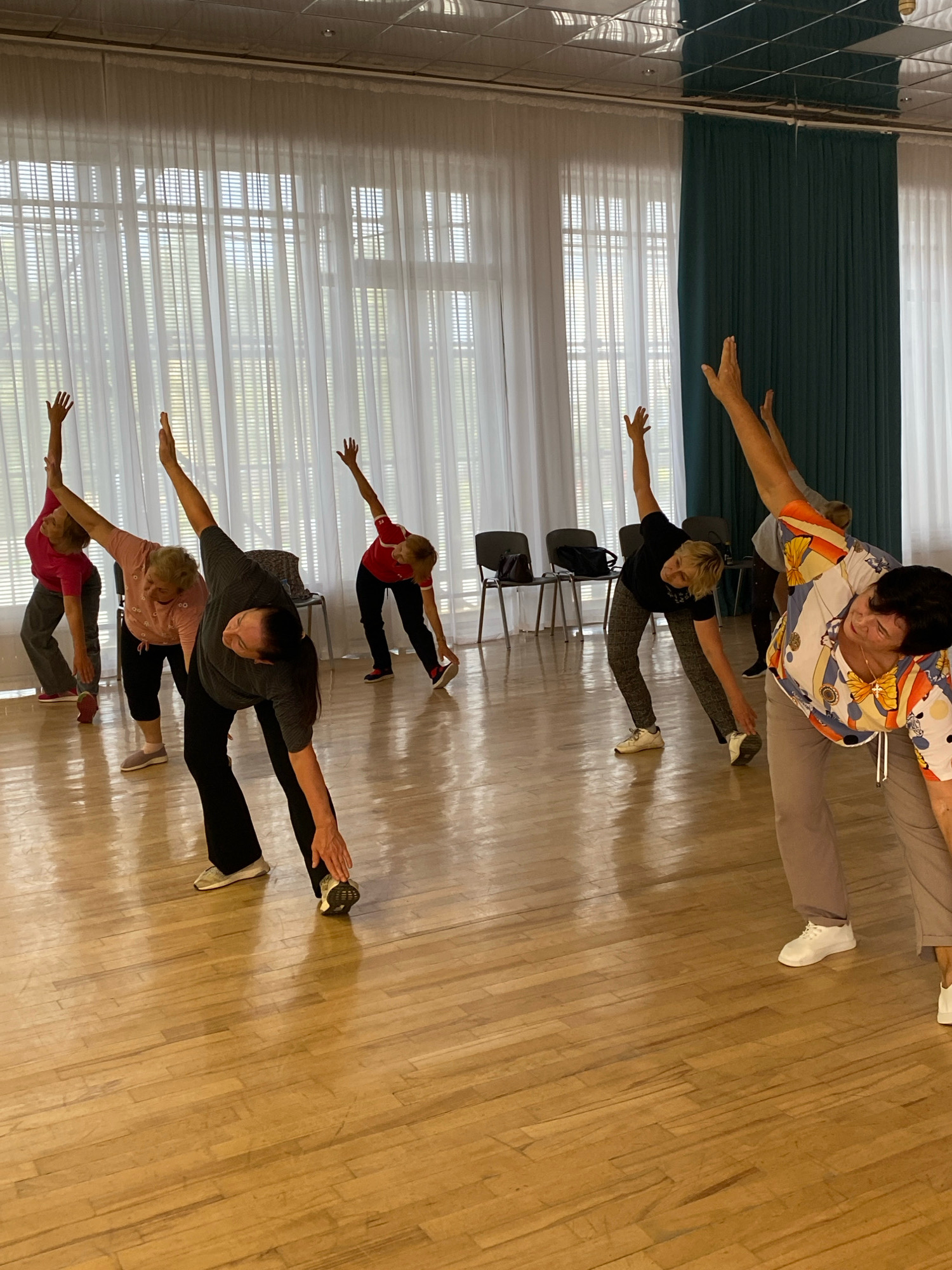 Seven individuals, all appearing to be adult females, are engaged in a stretching exercise in a room with a polished wooden floor. The room features large windows with white blinds and teal curtains. Several chairs are visible in the background. The people are positioned at varying distances from the camera, all in mid-stretch with their bodies bent sideways and arms extended upwards or downwards. The lighting is bright, suggesting an indoor setting during the daytime. No specific landmarks or distinguishing features of Ivanteyevka, Russia are discernible in the image.