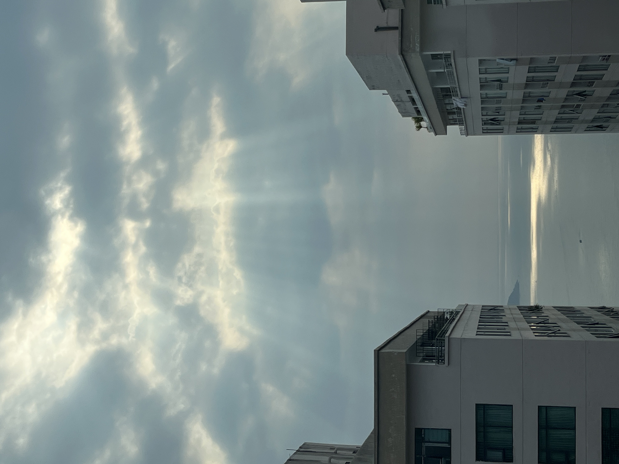 Captured from an elevated perspective in the city of Bac Nha Trang Ward, Vietnam, the image features a view between two multi-story buildings towards a body of water. The buildings are light-colored, with visible windows and balconies. Between the buildings, the sky is filled with clouds, and sun rays are breaking through. In the distance, a body of water reflects sunlight. A small boat can be seen in the water.