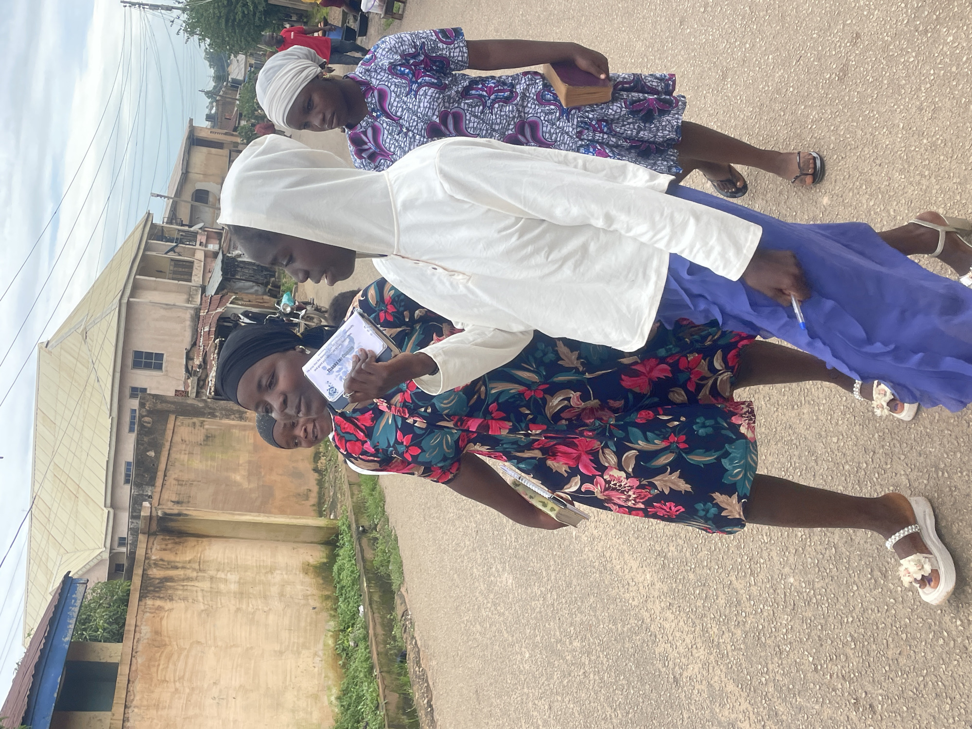 In the city of Jos, Nigeria, three young women walk on a paved road. The woman in the foreground, on the left, wears a dark blue dress with a vibrant floral print and white sandals adorned with flowers. She holds a book under her arm and a pamphlet in her right hand, which she is showing to the woman next to her. The woman in the middle wears a white hooded sweatshirt and a long, flowing blue skirt. She holds a pen in her right hand and a pamphlet in her left hand. The woman on the right wears a patterned dress and a headscarf. She holds a book under her arm. Behind them, a man in a red shirt and a person in a blue shirt are visible in the distance. Buildings line the street, with one building featuring a prominent beige facade and a blue-trimmed roof. The sky is overcast.