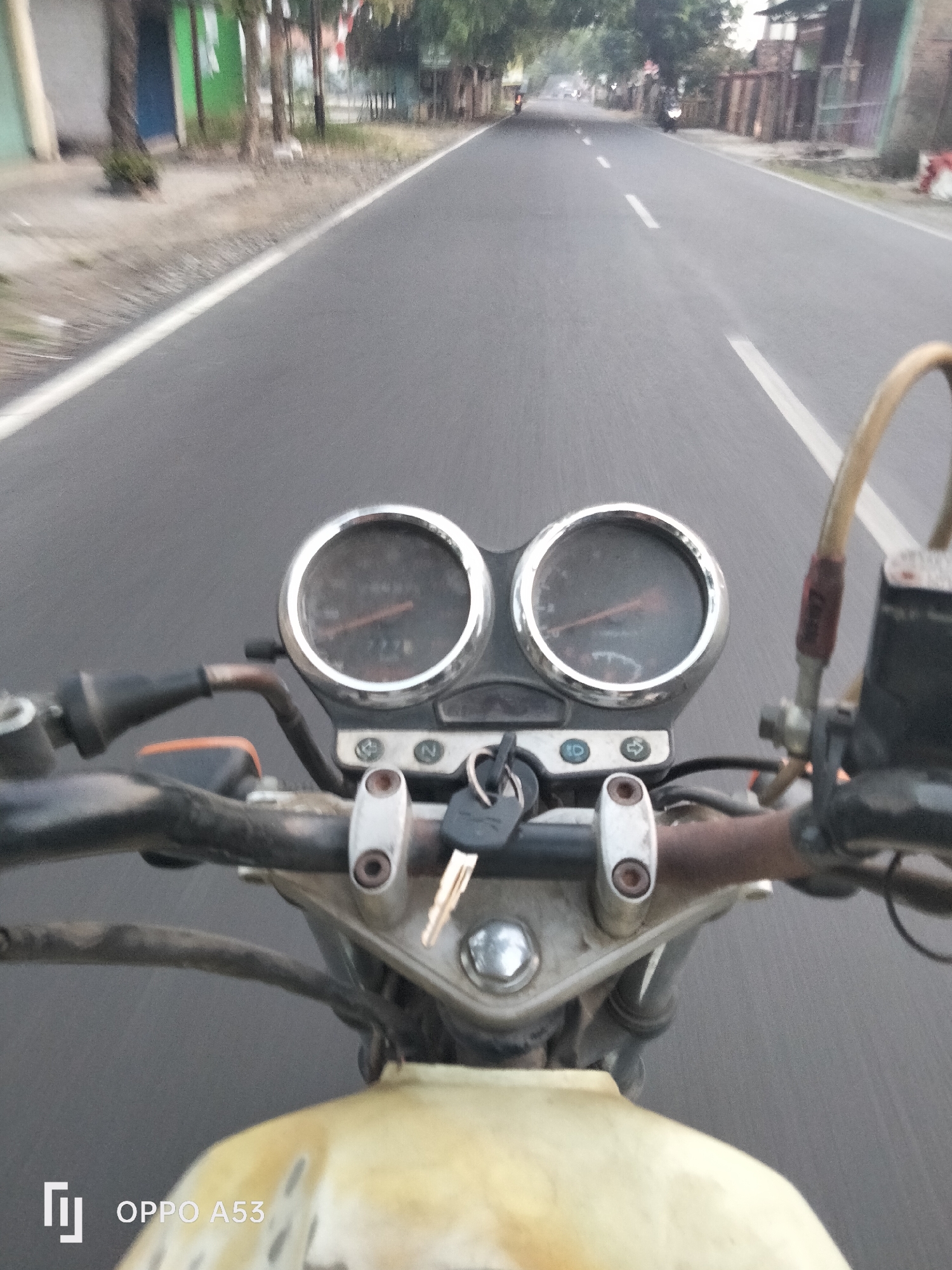 The image is a first-person perspective from a motorcycle traveling on a paved road in Karawang, Indonesia. The road is lined with sparse vegetation, trees, and buildings on either side, suggesting a semi-urban or rural setting. The sky appears overcast. The motorcycle's handlebars are visible in the foreground, with a chrome-rimmed speedometer and tachometer cluster. A set of keys is inserted into the ignition. The motorcycle's fuel tank is partially visible at the bottom of the frame, with the "OPPO A53" watermark in the lower left corner indicating the device used for capture. Several other motorcycles and vehicles are visible in the distance on the road.