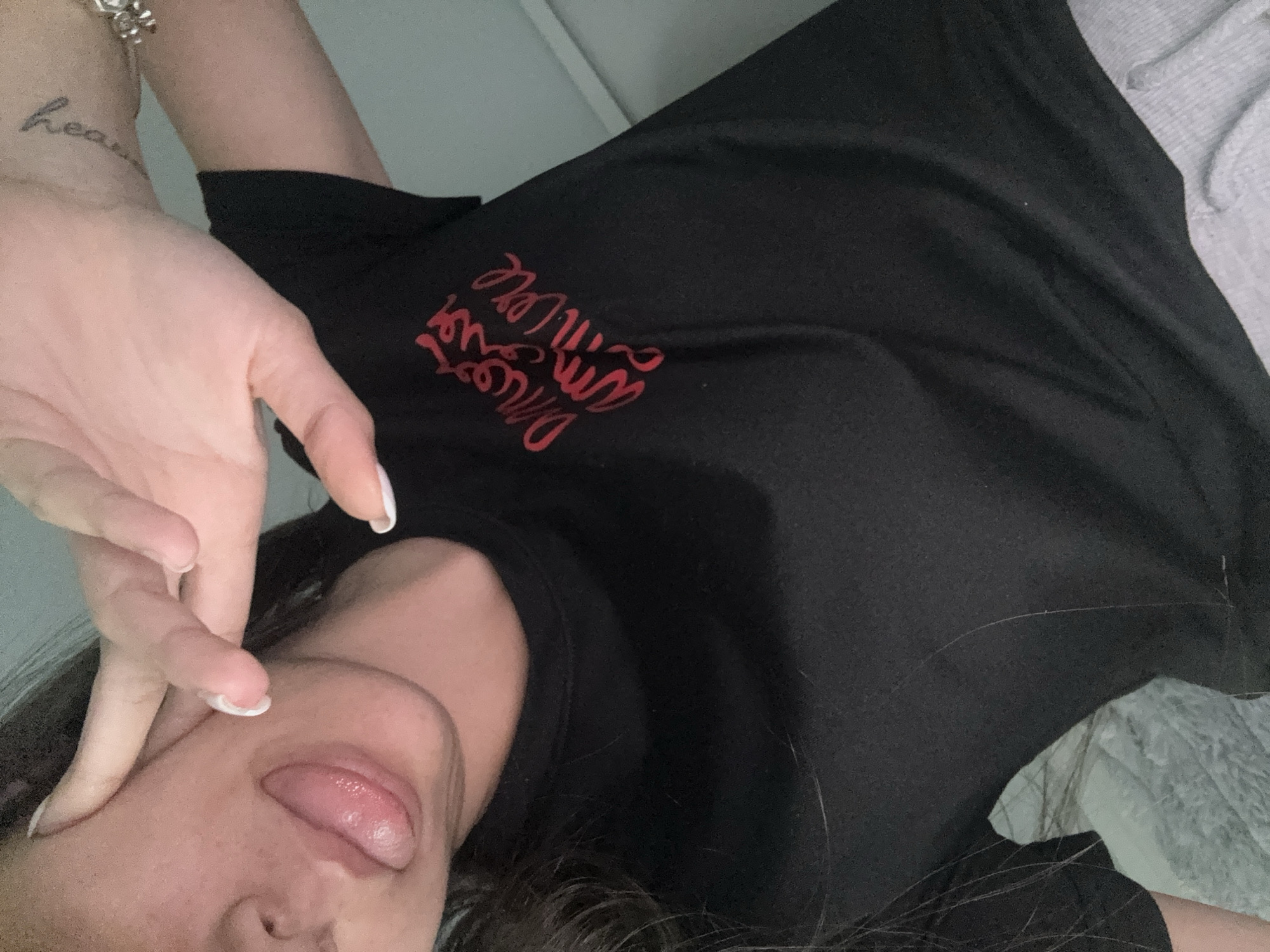 A woman lies on a light-colored surface. She is wearing a black T-shirt with red text. Her tongue is sticking out. Her left hand is raised towards her face. There is a tattoo on her wrist. A bracelet is visible on her wrist. There is no information that suggests that the image was taken in Sofia, Bulgaria.