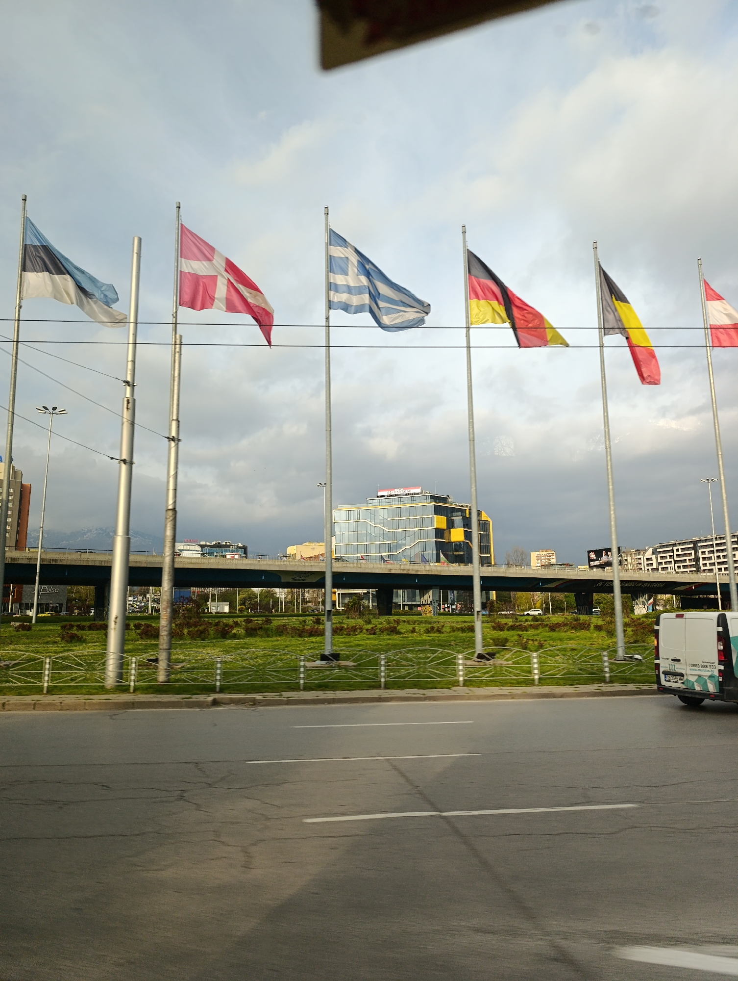 The image shows a street scene in Sofia, Bulgaria, with a row of flags representing different European countries fluttering in the wind. The flags from left to right are Estonia, Denmark, Greece, Germany, Belgium, and Austria. In the background, there is a highway overpass with modern office buildings visible beyond. A green strip of land runs along the road, separated by a low, decorative fence. A white van is partially visible on the right side of the image. The road itself is gray and marked with white lines. The sky above is overcast with gray clouds.