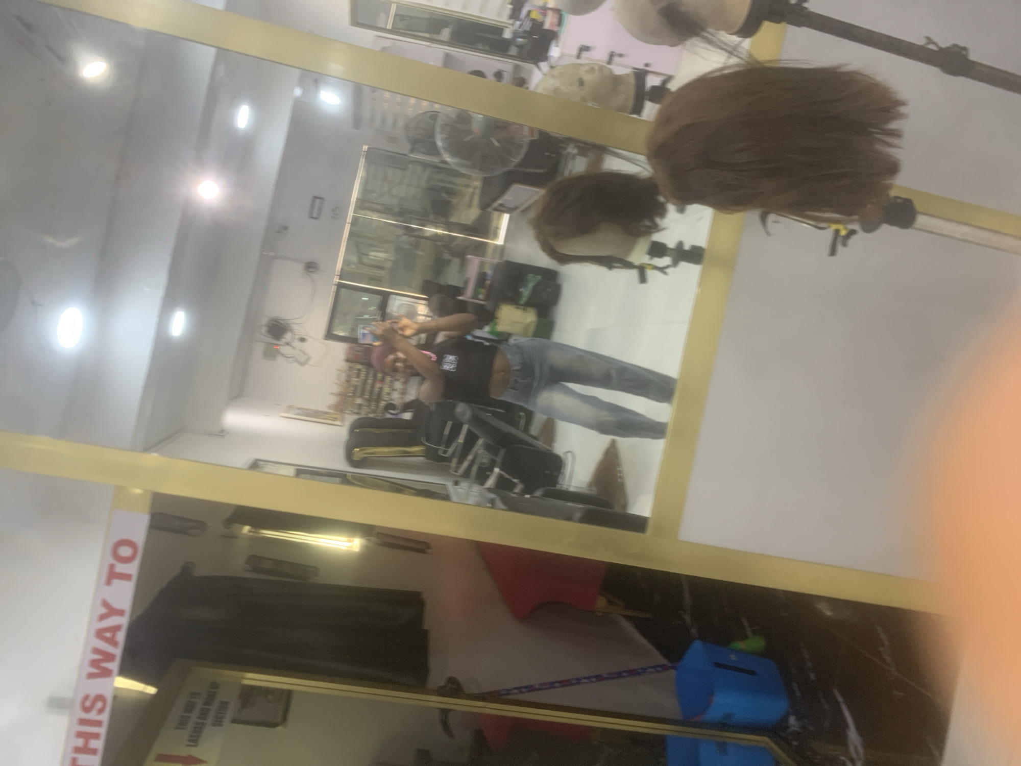 The image shows the reflection in a mirror of an interior space, likely a hair salon. A person wearing a black shirt and jeans is seen in the reflection, holding up a phone as if taking a picture. There are hair mannequin heads on stands in the foreground. Other hair salon equipment, such as chairs and mirrors, is visible in the background, along with ceiling lights. There's a sign on the side of the mirror in the foreground that reads "THIS WAY TO". It's difficult to determine the precise location in Unknown, Unknown due to the lack of identifiable location markers.