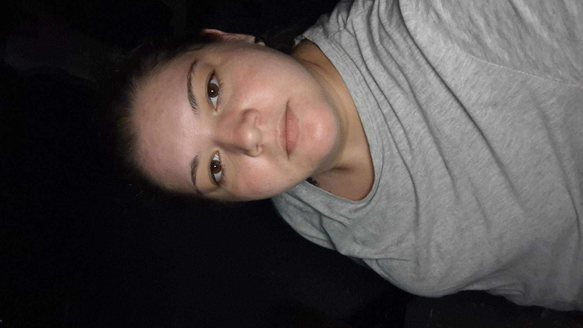 The image is a close-up selfie of a woman with brown eyes, fair skin, and brown hair pulled back. She is wearing a gray t-shirt. The background is dark and out of focus, suggesting it might be nighttime or a dimly lit indoor environment. There are no discernible landmarks or contextual clues within the image that would indicate a specific location in the city of Shumen, Bulgaria. The scene is static, with the subject looking directly at the camera.