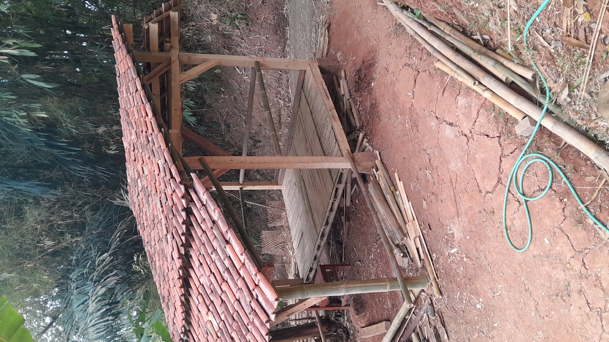 The image depicts an unfinished bamboo structure with a tiled roof in Sumedang, Indonesia. The structure appears to be a simple open-air pavilion or shelter. The roof is made of red clay tiles. The floor of the structure is constructed from horizontally laid bamboo poles. Wooden posts support the roof. Several bamboo poles are piled on the ground around the structure, suggesting that it is under construction or has recently been built. A green hose is coiled on the red, dry, cracked earth in the foreground. The background shows dense foliage, likely bamboo and other tropical plants, and a hillside. The overall scene suggests a rural or semi-rural setting within the city of Sumedang.
