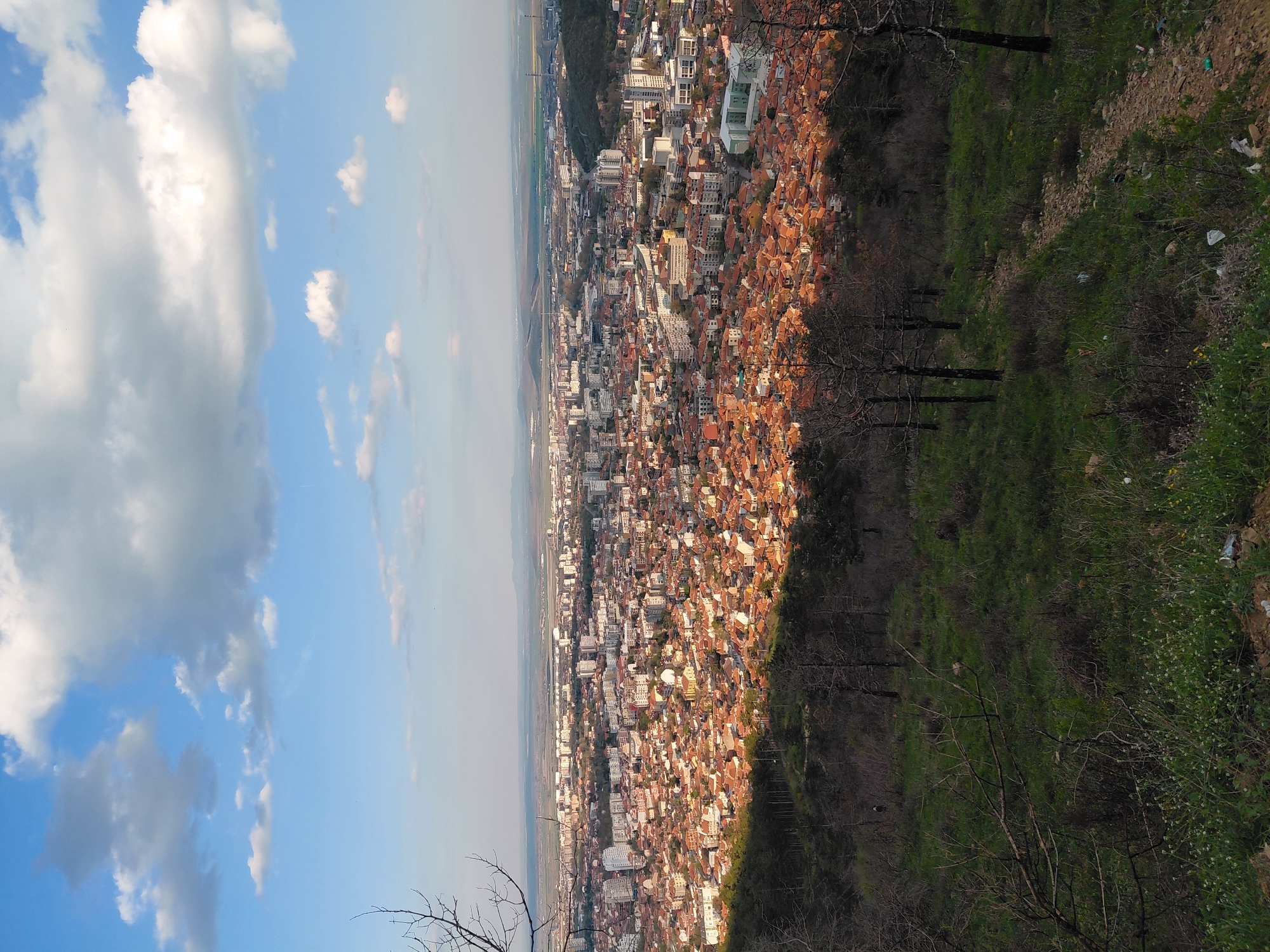 The image shows an aerial view of Sliven, Bulgaria, with a blue sky and scattered clouds in the upper left. The city extends along a valley, characterized by densely packed buildings with predominantly orange and white roofs. A green, vegetated hillside borders the urban area on the right side of the frame. Sparse trees are visible on the hillside, suggesting possible forestation. The landscape stretches into the distance, with hazy mountains or hills forming the horizon. There is no evidence of human presence or activity within the frame. The photograph appears to have been taken from a high vantage point, offering a panoramic perspective of the city and its surrounding environment.