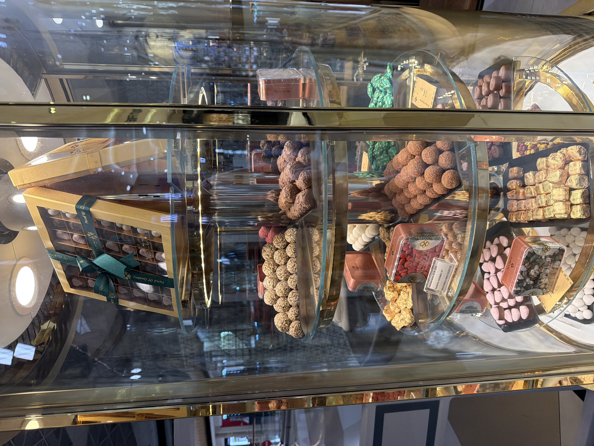 The image shows a cylindrical glass display case filled with an assortment of chocolates and candies. The display case has gold-colored trim and multiple glass shelves. On the top shelf, a large box of chocolates with a green ribbon is prominently displayed. Below this, various trays hold different types of chocolates and candies, including round truffles, small chocolate bars, and candies in clear plastic boxes. Some of the candies appear to be fruit-flavored, such as red and pink berry-shaped candies. The chocolates are arranged in neat rows and piles. The background is blurred, suggesting an indoor environment like a store or market. There are no people visible in the image. The location of Khazar Raion, Azerbaijan is not determinable from the image content.