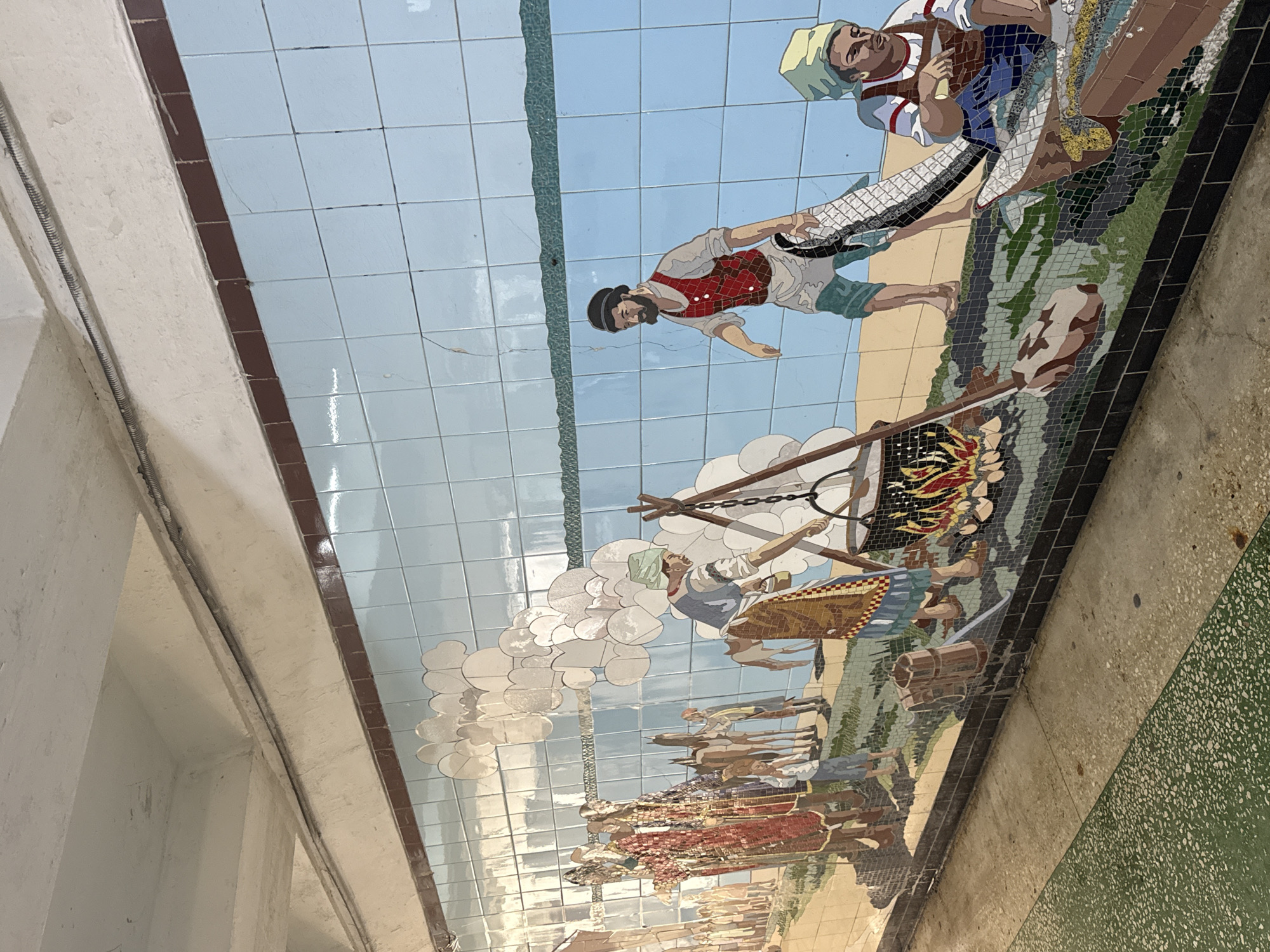 A tile mosaic in Rostov-on-Don, Russia, depicts various figures engaged in labor and social activities against a backdrop of blue sky and landscape. The mosaic shows groups of people working with tools around a fire, carrying objects, and processing materials, potentially depicting industrial or agricultural scenes. The mosaic is tiled on a wall in what appears to be a tunnel, passageway, or underground area, as indicated by the adjacent architectural features.