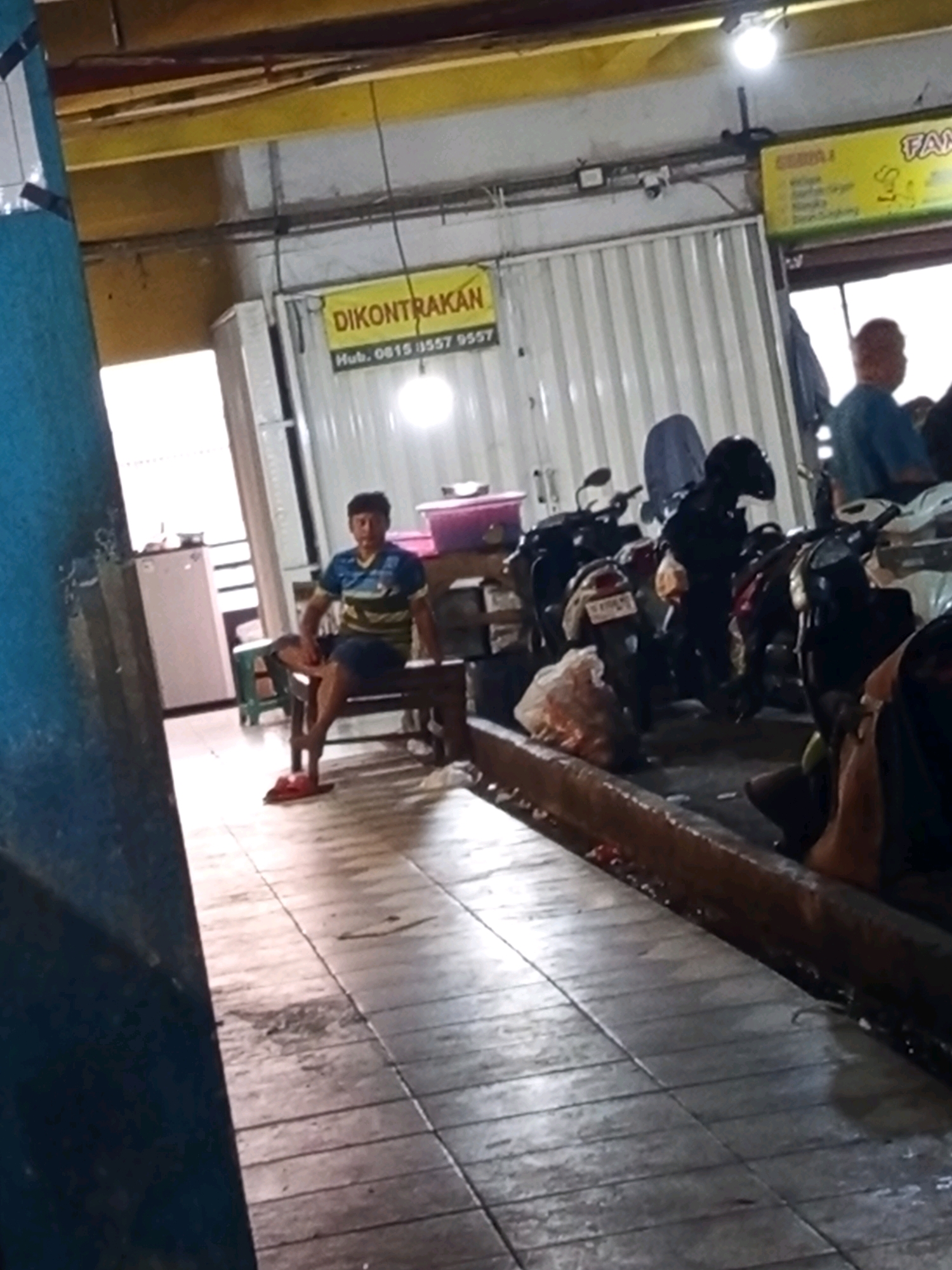 The image depicts a dimly lit indoor scene, likely a market or workshop, with a focus on motorcycles parked in a row. A young man, wearing a striped t-shirt and dark shorts, is seated on a wooden bench in the foreground, facing left. His posture suggests relaxation or waiting. To the left of the man, a white refrigerator is visible. In the background, several motorcycles are parked, some with helmets placed on them. A yellow sign with Indonesian text, "DIKONTRAKAN," which translates to "For Rent," is prominently displayed on a white corrugated wall. Another yellow sign with text "FAM" is partially visible to the right. The floor appears to be tiled and wet in some areas, possibly indicating recent cleaning or a spill. The overall environment suggests a functional, possibly commercial space, with elements of Indonesian street life.