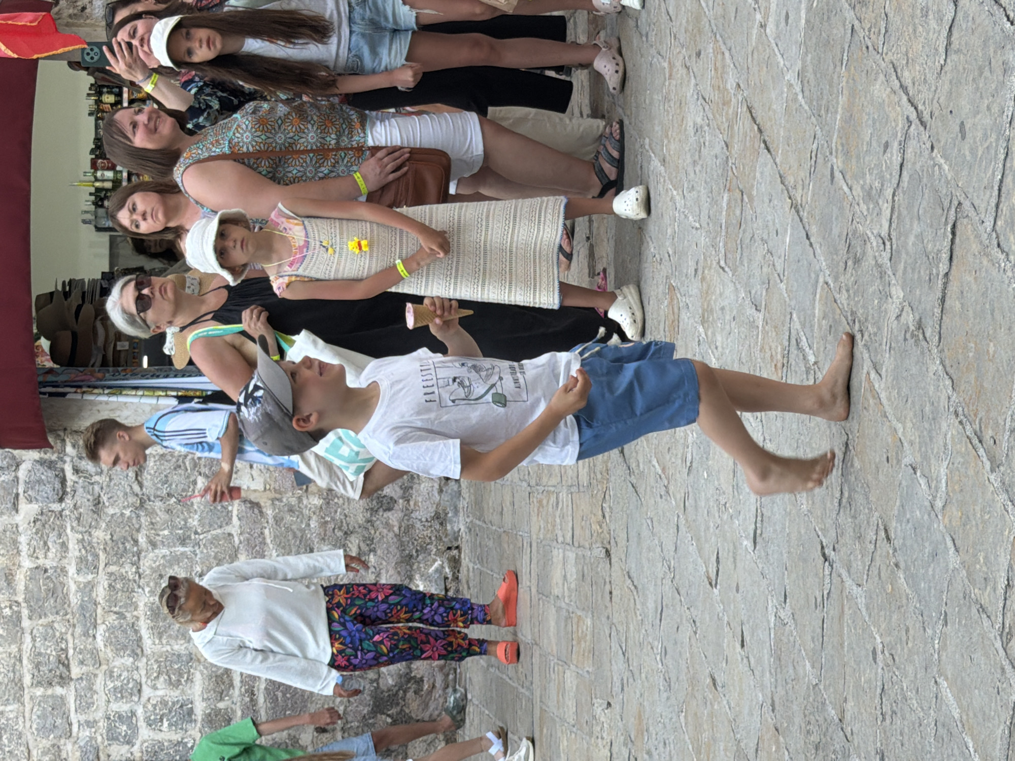 The image depicts a street scene in Budva, Montenegro, with a focus on a young boy in the foreground wearing a white t-shirt, blue shorts, and a baseball cap. He is walking barefoot on a stone-paved street. Behind him, a group of people are visible, including a woman in a white hoodie and floral pants, a young man in an Argentine football jersey holding a drink, and several other individuals. The background shows a stone wall and the entrance to a shop with various items displayed, including hats and bottles. The overall atmosphere suggests a casual outdoor environment, likely a pedestrian area within the city.