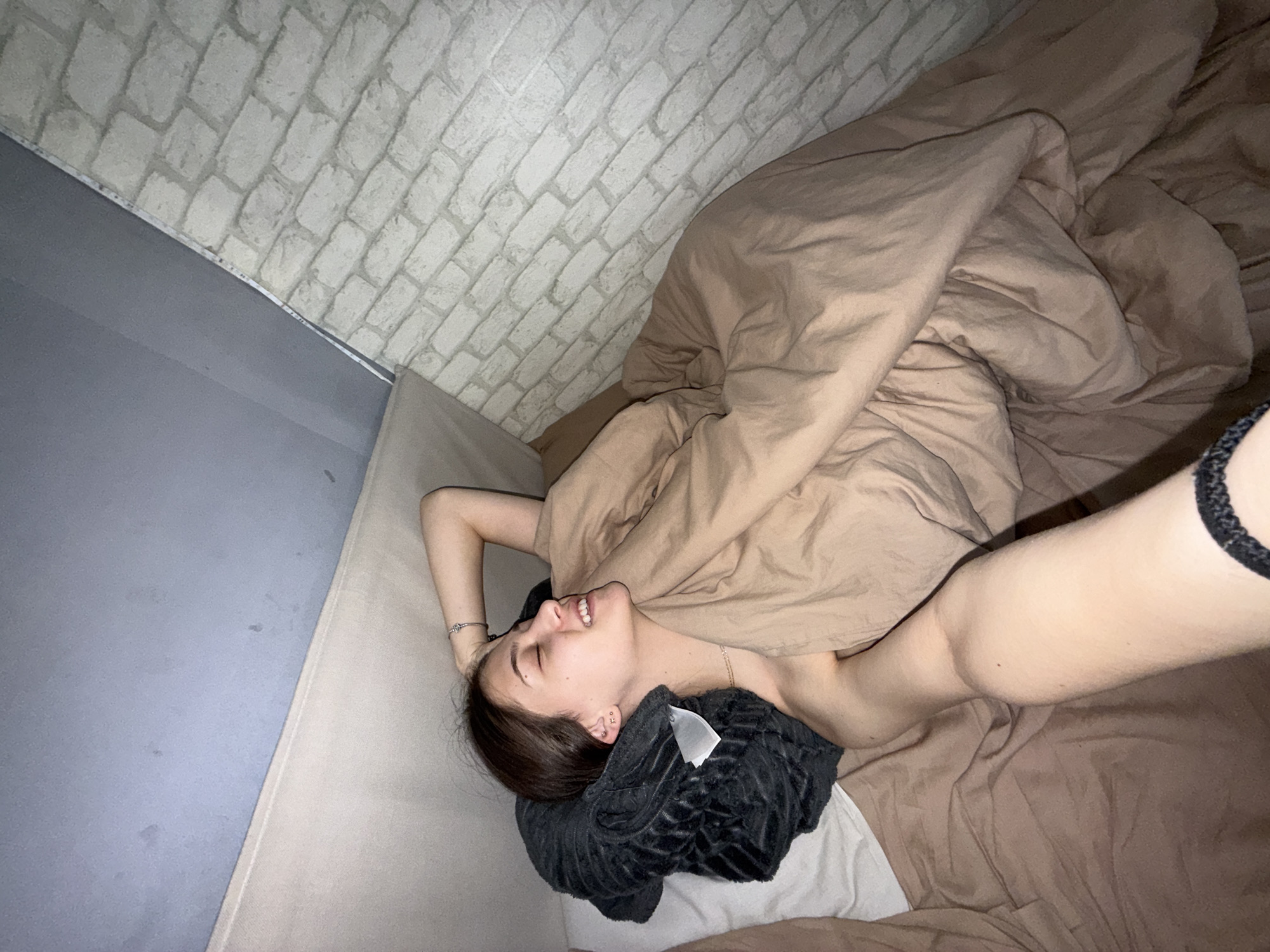 A young woman is lying in bed, facing upwards and slightly to the left of the frame. She is covered in a brown duvet and has a gray pillow under her head. Her right arm is raised towards the camera, revealing a bracelet. The bedroom has a wall with a white brick pattern on the left and above. There is no information in the image to indicate that the image was taken in Sofia, Bulgaria.