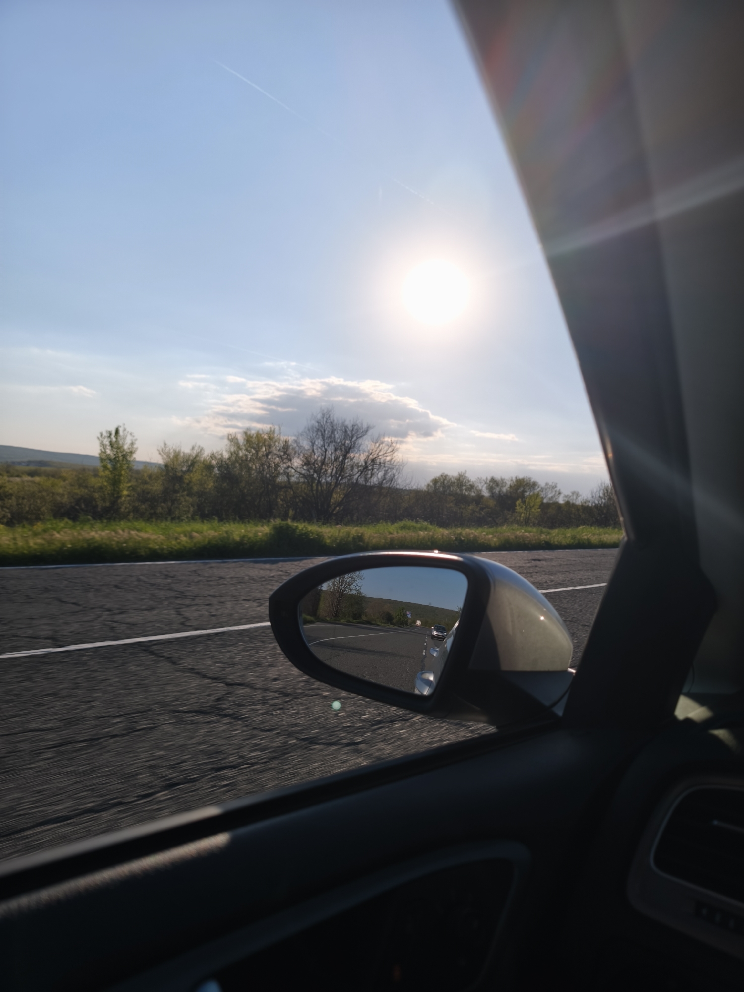 The image shows a view from inside a moving car. The car is driving on an asphalt road with a white lane marker. A side mirror is visible reflecting the road behind the car, including another vehicle. Outside the car window, there is a landscape of green fields and trees under a bright sky with the sun shining. The image was taken in Rudnik, Bulgaria.