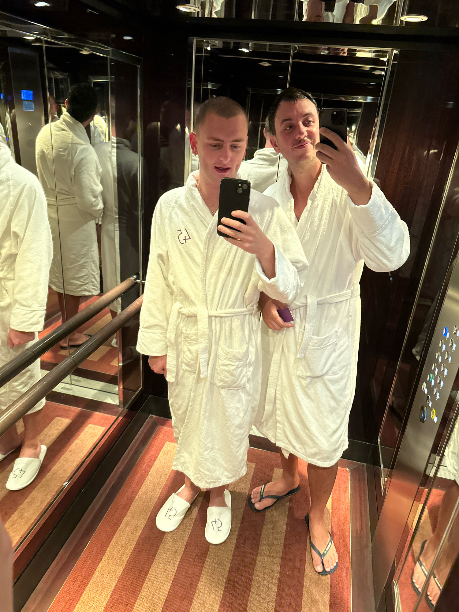 Two individuals are captured in a mirror selfie taken inside an elevator. Both are wearing white terrycloth bathrobes and slippers. The individual on the left has "45" embroidered on their slippers, while the individual on the right has "12" on theirs. The person on the right is holding a purple object. The elevator has mirrored walls and a wood-paneled ceiling. The floor of the elevator is carpeted with alternating stripes of orange and brown. The elevator buttons are visible on the right-hand wall. The scene does not provide any specific visual cues to identify the location as London, United Kingdom.