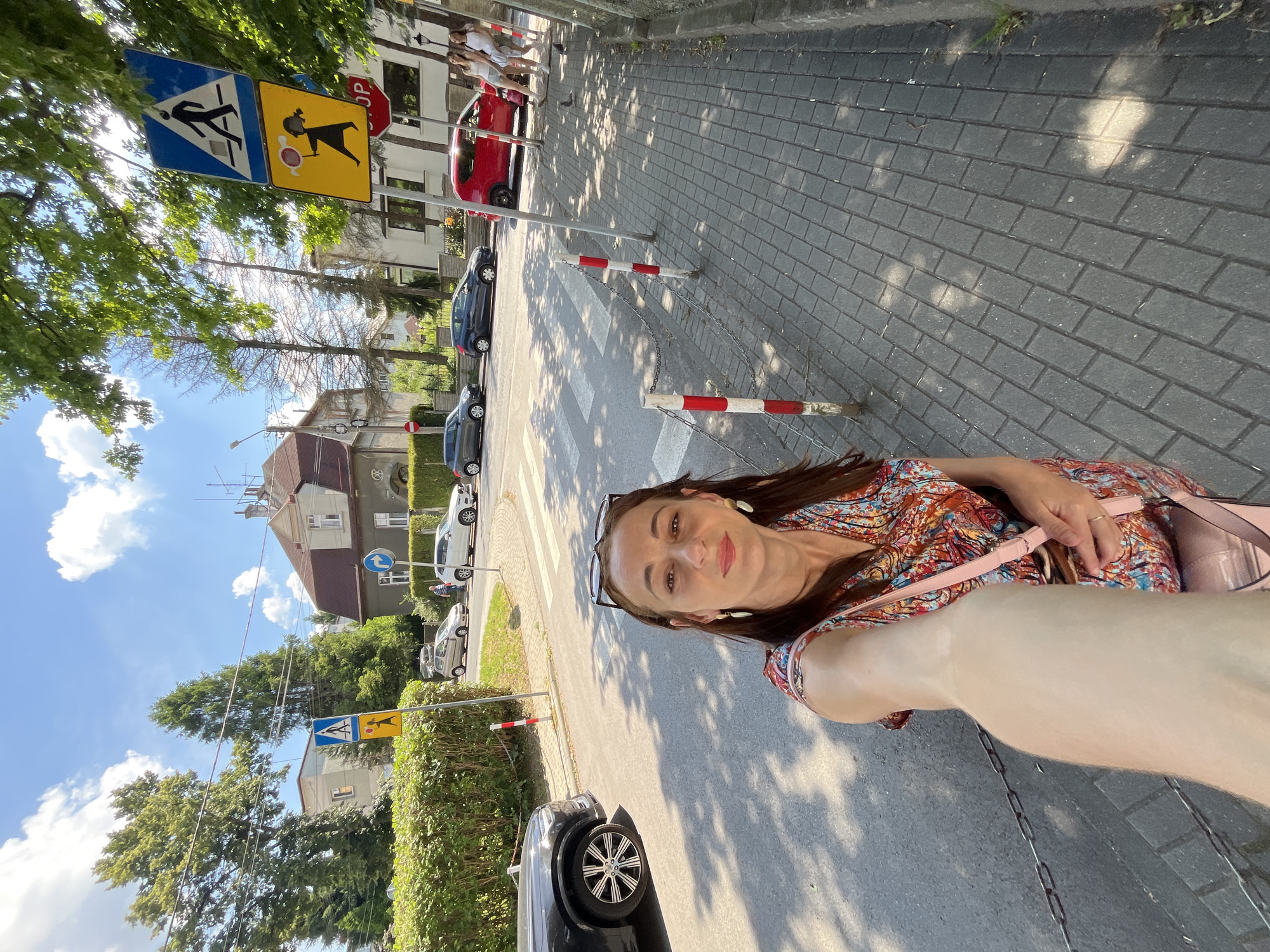 The image shows a woman taking a selfie outdoors. She is wearing a colorful patterned dress and glasses. A pink purse is slung over her shoulder. The scene appears to be a street or sidewalk with a crosswalk. There are cars, buildings, trees, and a blue sky with clouds visible in the background. Two traffic signs are also present; one is blue and white with a pedestrian symbol, and the other is yellow with a symbol of a person pushing a stroller. The time of day appears to be daytime, as indicated by the bright sunlight and shadows. The woman has a slight smile on her face, suggesting a pleasant mood. The ground is paved with cobblestones.