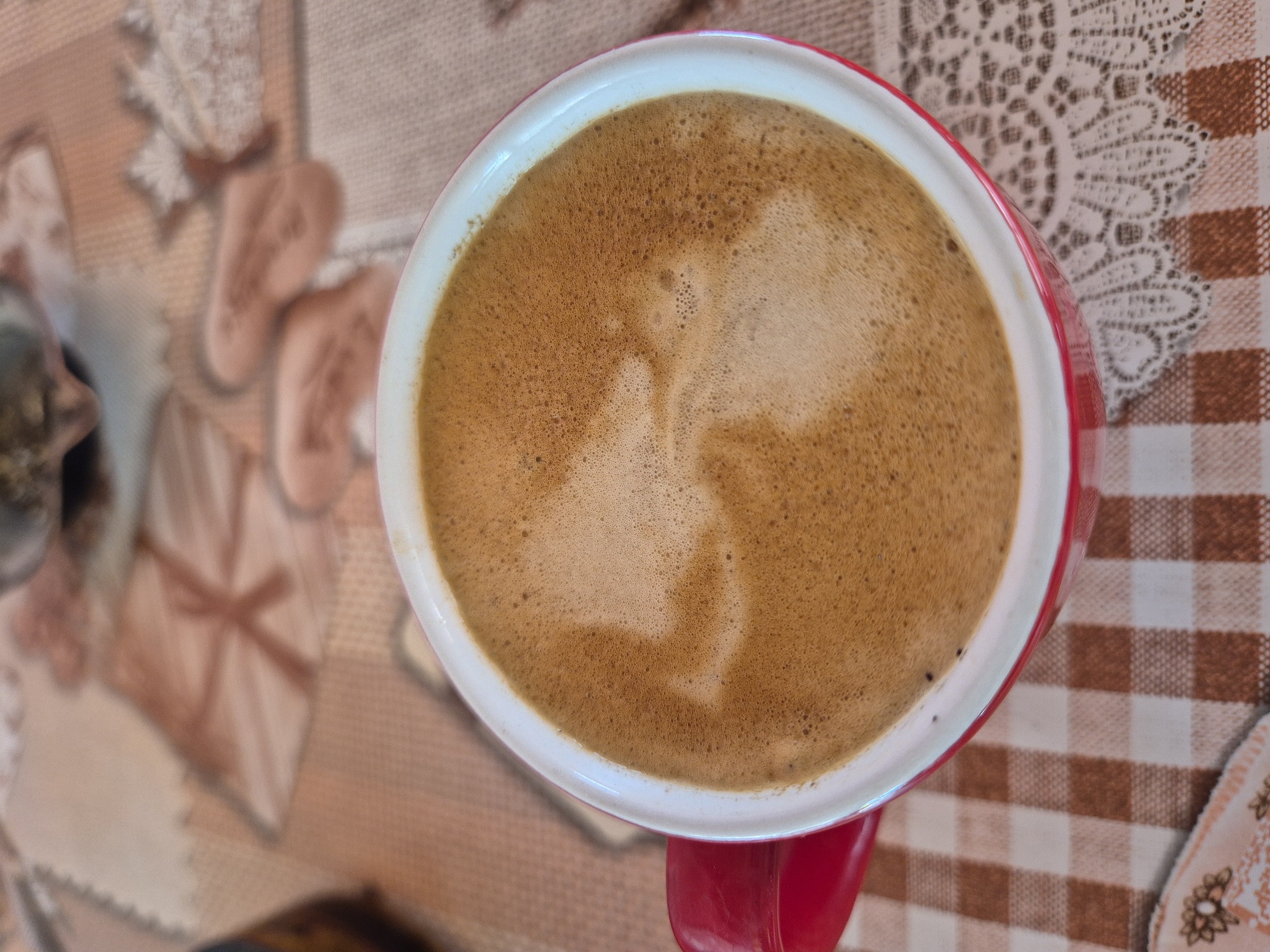 A close-up, overhead shot captures a red mug filled with frothy coffee, resting on a checkered tablecloth. The coffee's surface displays a light brown, foamy texture with some darker specks visible, suggesting it's freshly brewed. The mug has a white rim and a bright red exterior, with its handle also visible. The tablecloth features a brown and white checkered pattern, with a delicate lace trim along one edge. In the background, out of focus, a larger tablecloth or placemat with an abstract floral or geometric pattern is visible. The overall impression is one of a casual, domestic setting, possibly a breakfast or coffee break.
