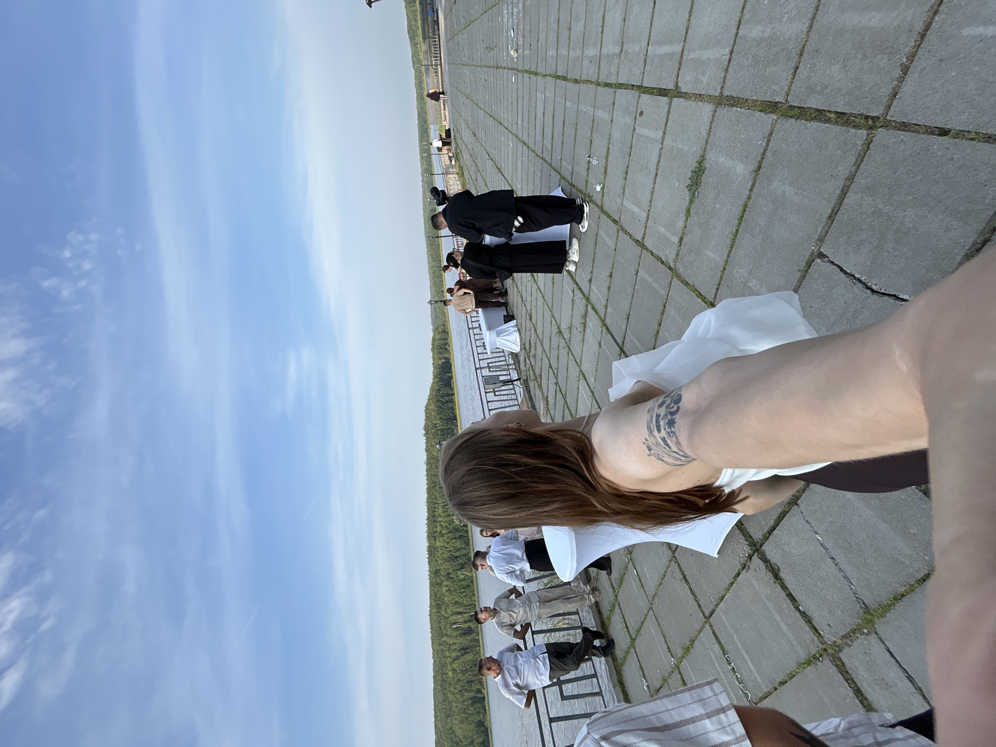 The image depicts an outdoor event taking place on a paved area overlooking a body of water, likely the Kama River in Chaykovsky, Russia. Several individuals are present, some standing near a railing by the water, while others are gathered around tables draped with white cloths. One person in the foreground is taking a selfie, with their arm and shoulder visible. The attire of the attendees suggests a semi-formal gathering or celebration. The environment is characterized by a clear sky with scattered clouds and a tree-lined shore in the background. The paved ground is made of large concrete slabs with grass growing in the cracks.